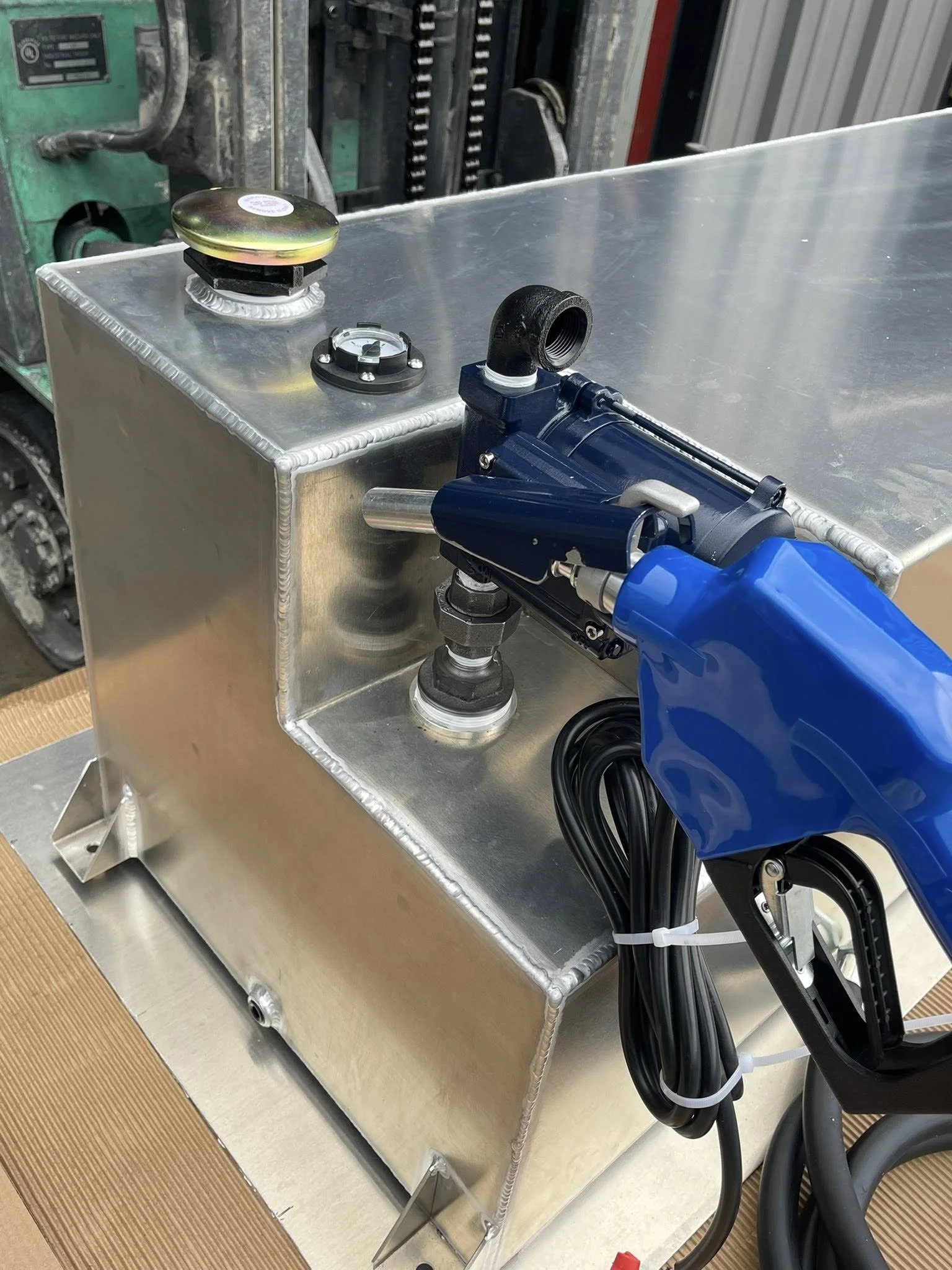 Close-up of a metallic tank with a pressure gauge, a capped inlet, and a blue pneumatic tool connected with black hoses, in an industrial setting.