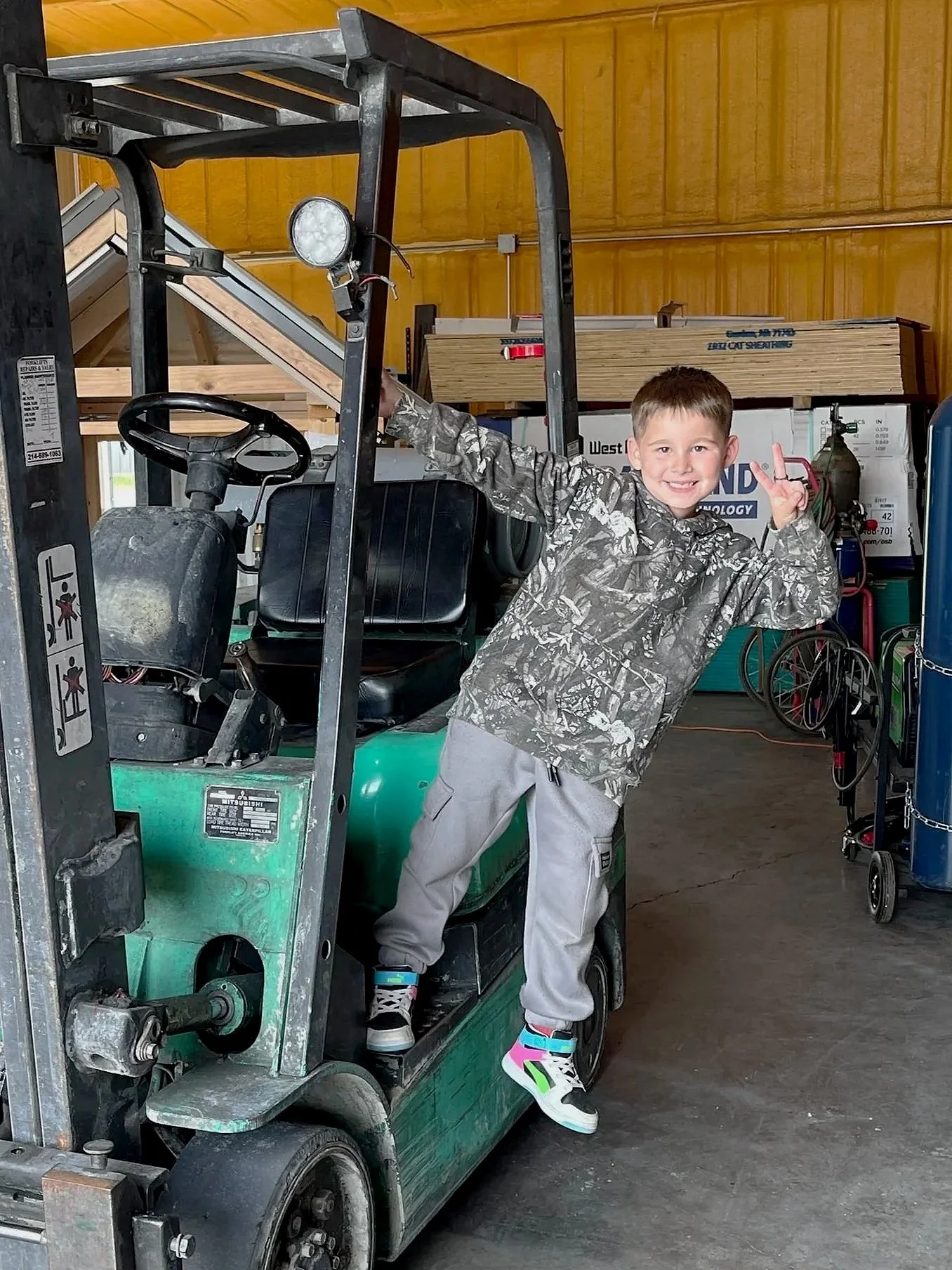 A boy in gray sweatpants, a camouflage jacket, and colorful sneakers is smiling and posing while holding onto a green forklift inside a warehouse with a wooden ceiling and shelves filled with boxes and tools in the background.