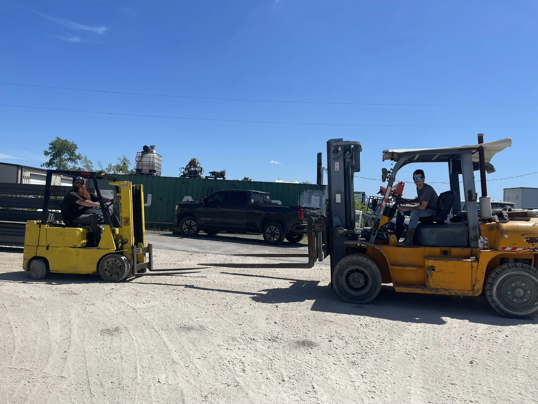 Two forklifts on a gravel surface, with people operating them. One forklift is yellow with a person wearing a mask and glasses, the other is rusty yellow with a person smiling and wearing sunglasses. There are trucks and containers welding