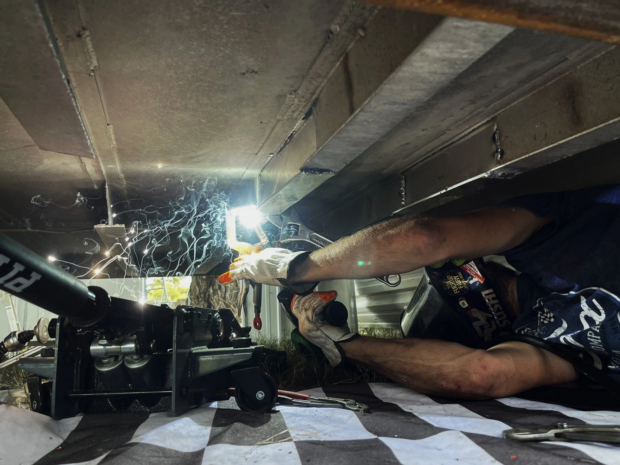 A person welding underneath a metal structure, creating sparks and bright light, wearing gloves and a welding helmet.