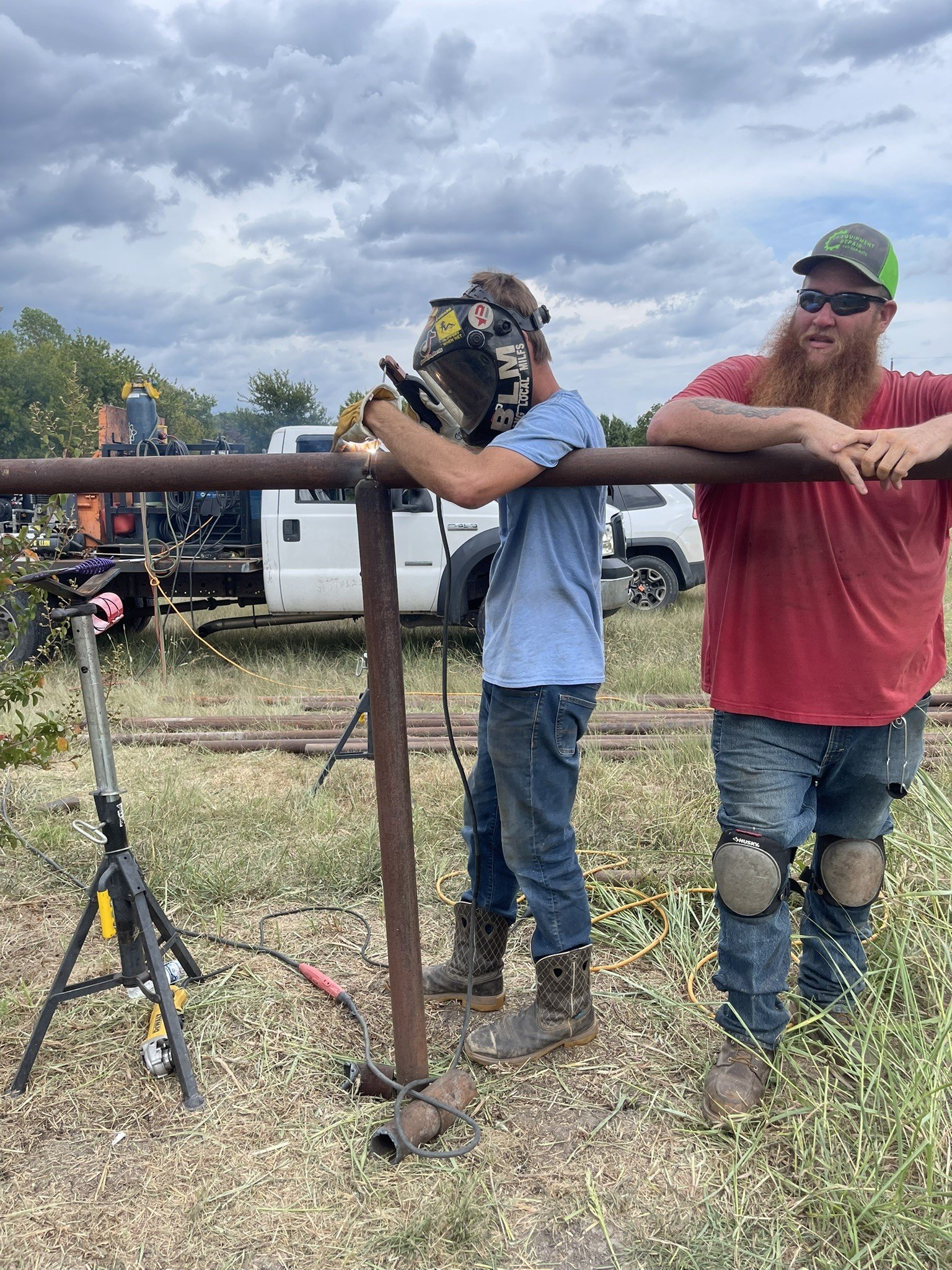 Two men working outdoors on a metal pipe, with a welding tool. One man is welding while the other watches, with cars and cloudy sky in background.