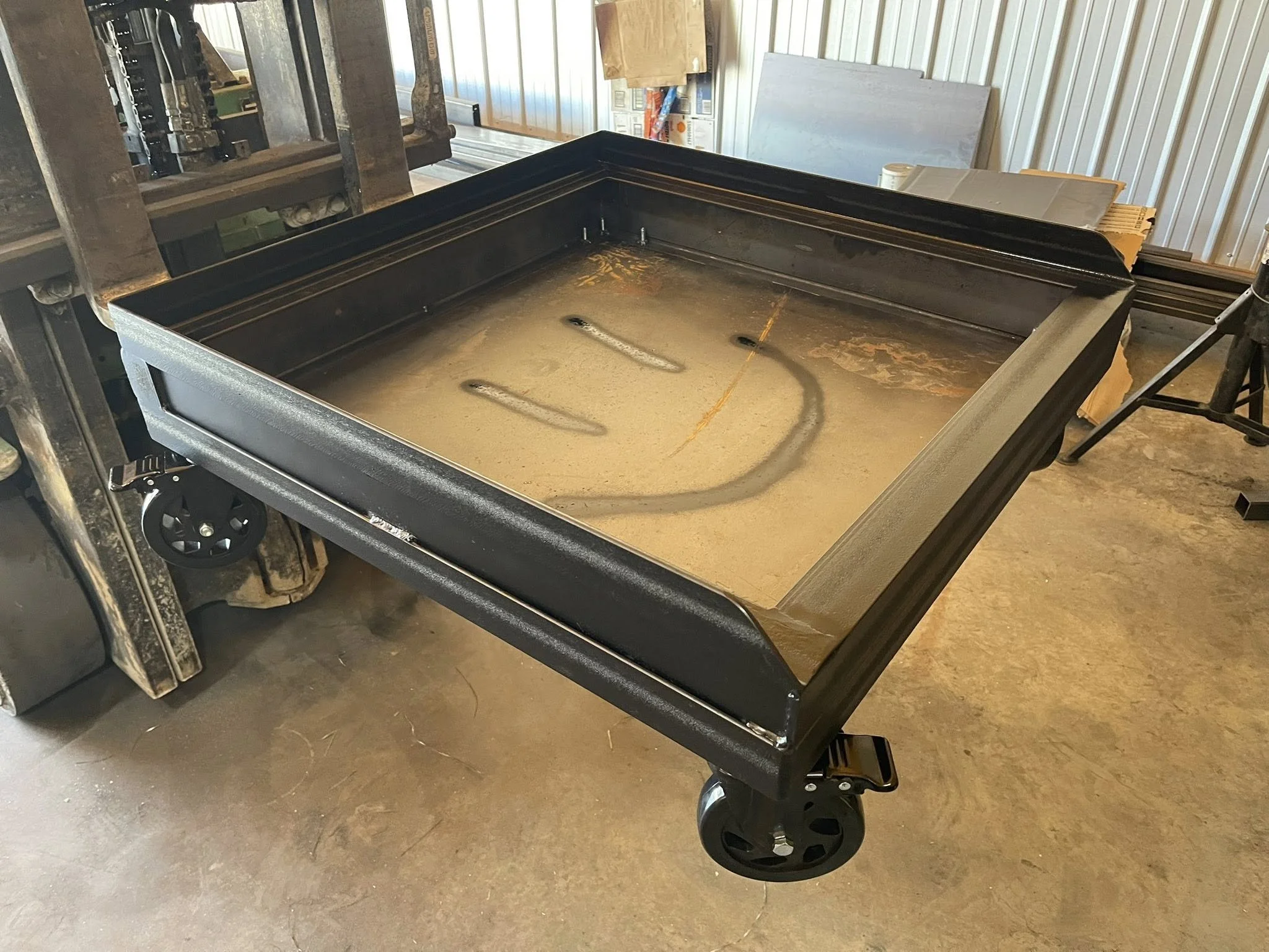 A black travel cooler or storage chest with wheels on the bottom, located in a workshop or garage setting with metal walls, cardboard boxes, and assorted materials in the background.