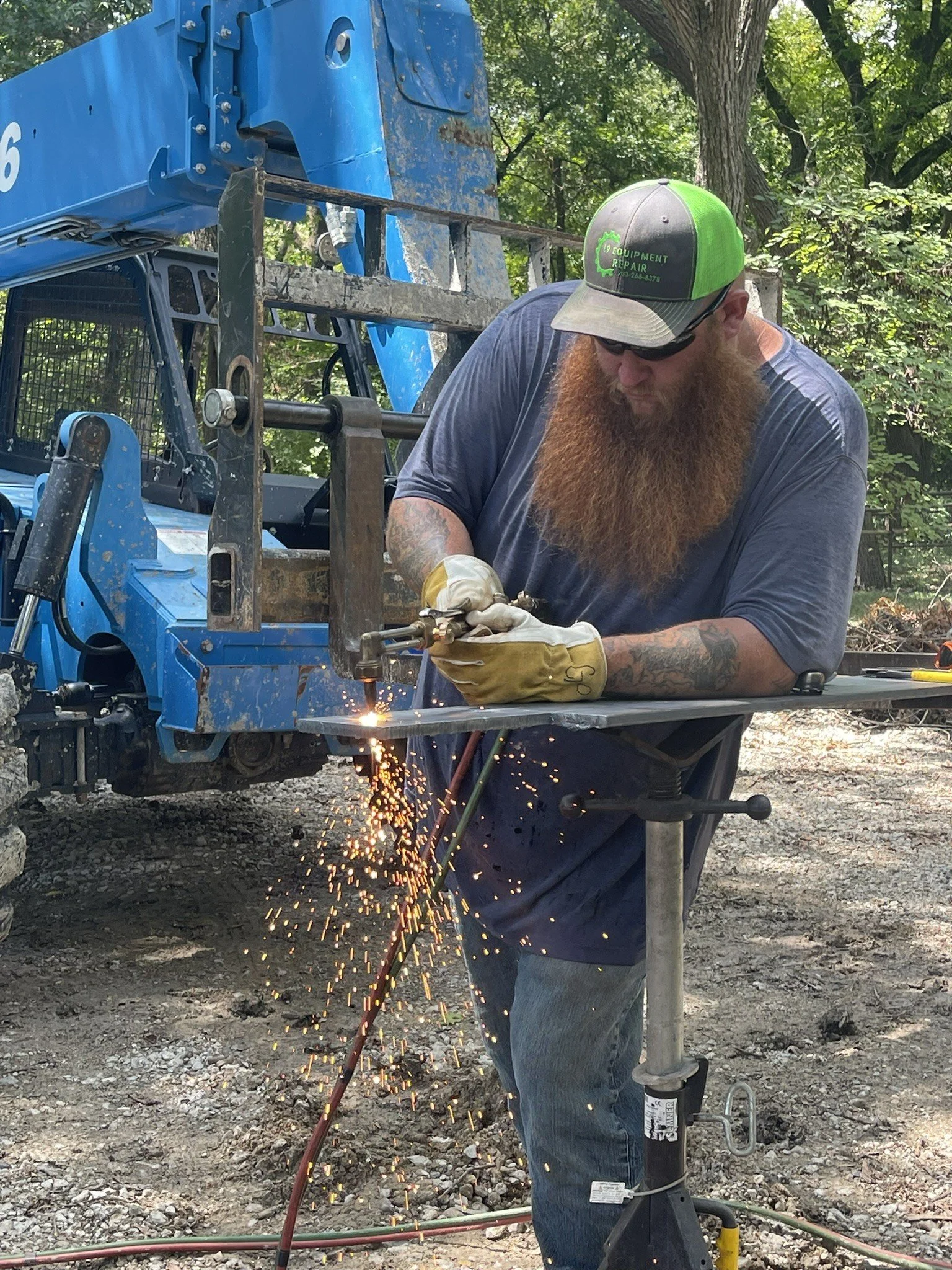 Man with a long beard wearing glasses, a green and gray cap, and protective gloves welding metal outdoors near construction equipment.