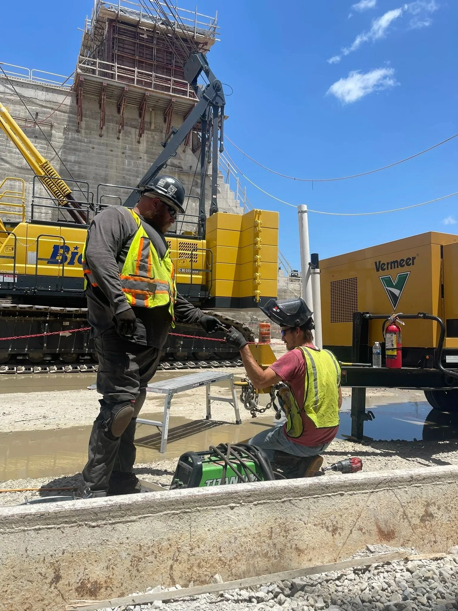 Two construction workers, wearing safety helmets and vests, are working near railroad tracks at a construction site. One worker is standing, and the other is kneeling, shaking hands. There is construction equipment, including a yellow crane and generator, in the background, under a clear blue sky.