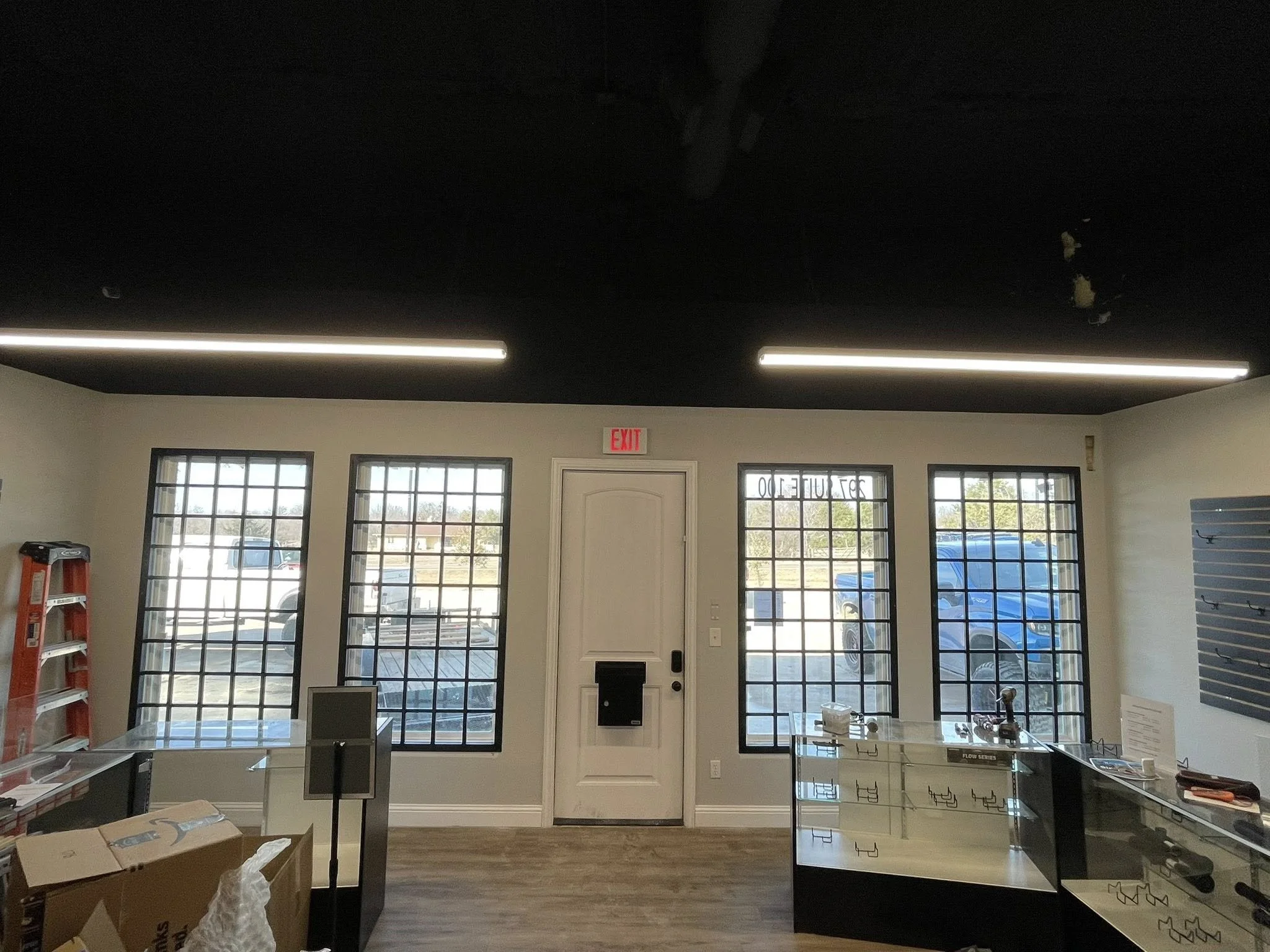 Interior of a commercial or office space with high ceilings, large windows, and modern lighting fixtures. There are some cardboard boxes and office equipment on the counter and floor, and a red ladder is visible on the left side of the image.
