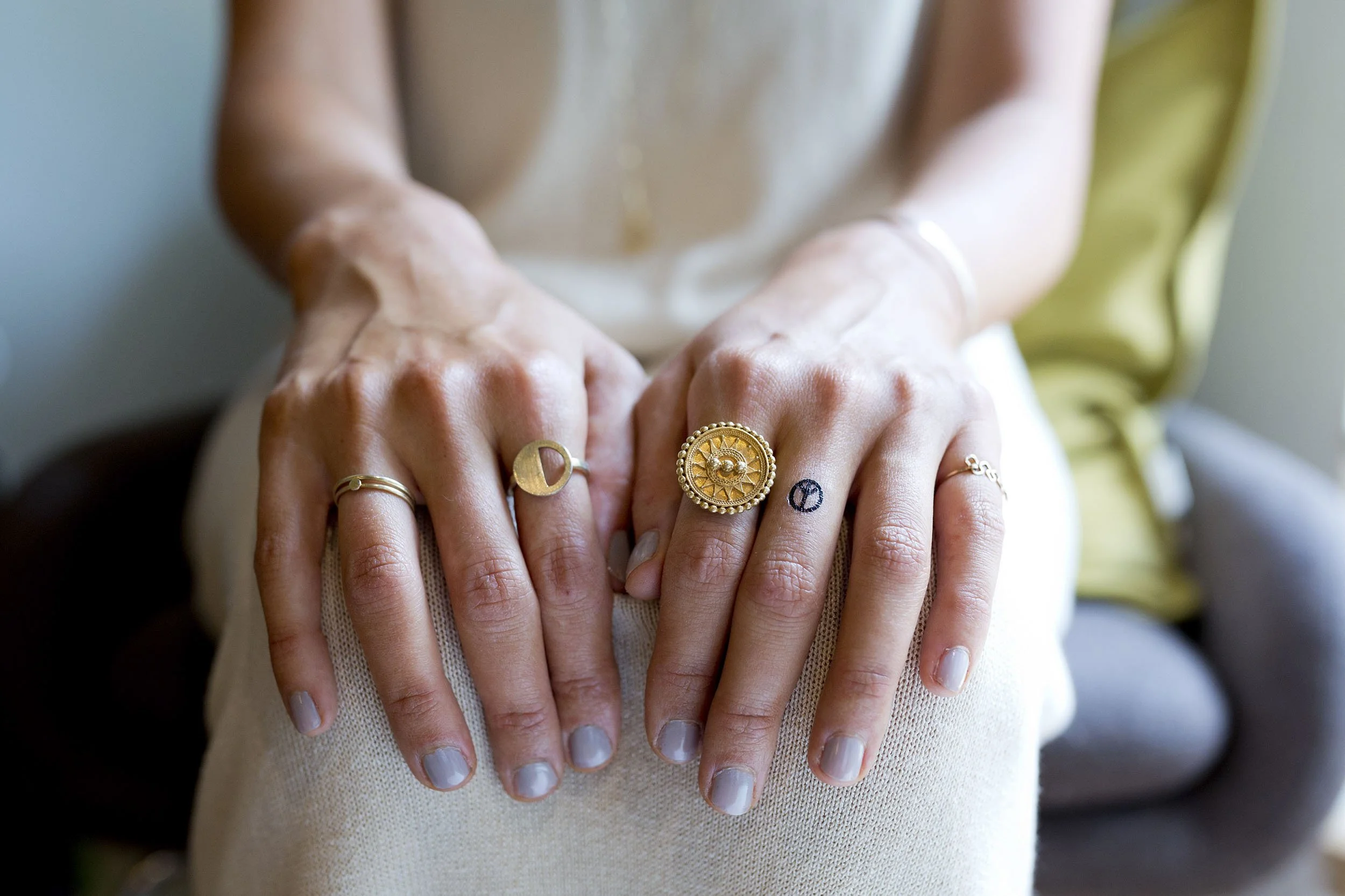 Nahaufnahme von Händen mit verschiedenen Ringen, auf einem Bein ruhend, im Hintergrund ein Sitzbereich.