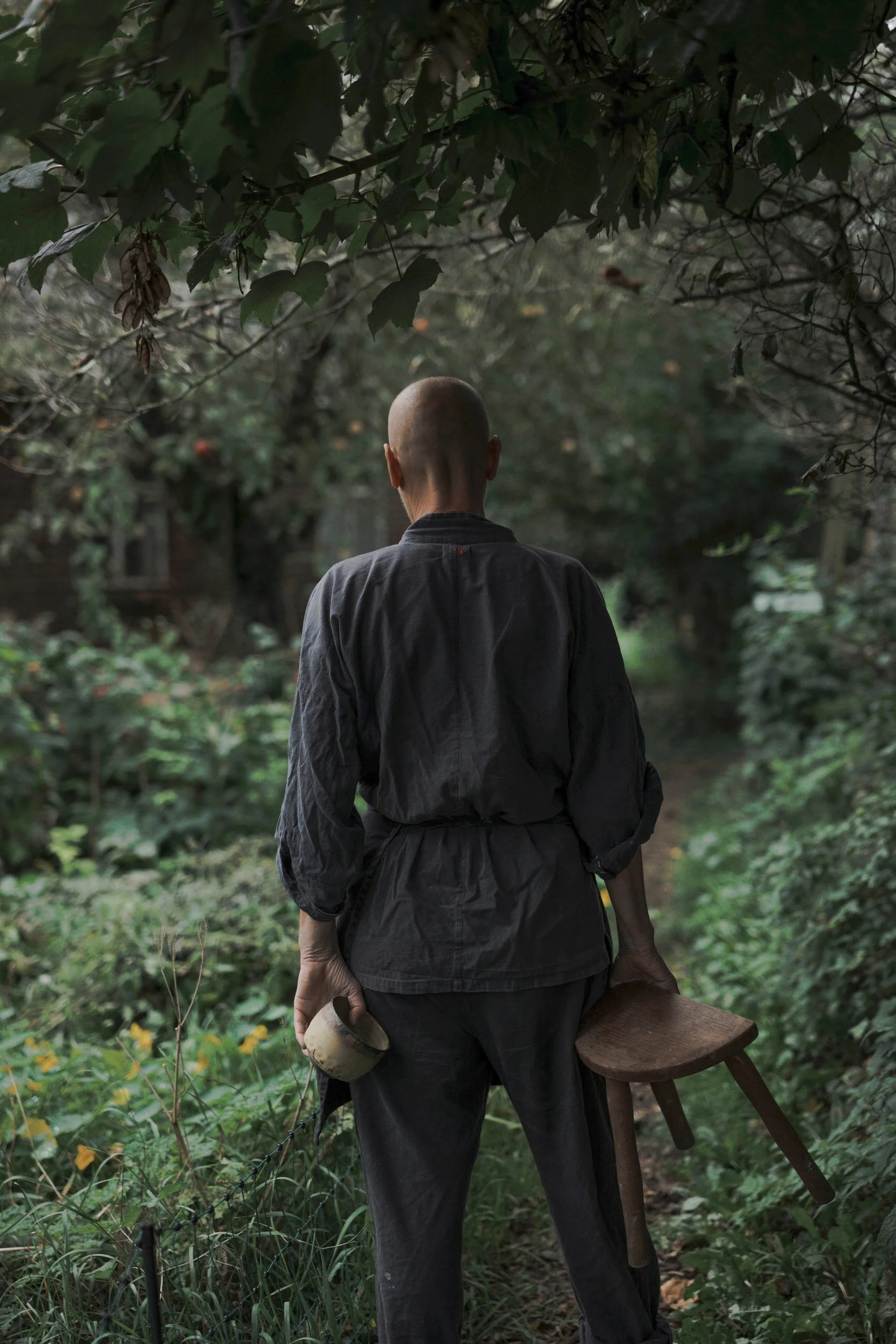 Eine Person mit kurzen Haaren, die von hinten gezeigt wird und eine schwarze Jacke trägt, steht in einem grünen Garten. Sie hält eine Keramik in der rechten Hand . Um sie sind zahlreiche grüne Pflanzen und Bäume sichtbar.