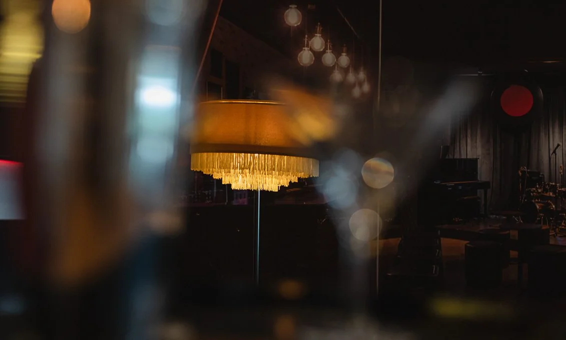 Ein dunkler Raum mit Bühnenmusik-Ausrüstung, einer Stehlampe mit goldener Fransen- schirm und einer Reihe von Glühlampen an der Decke.