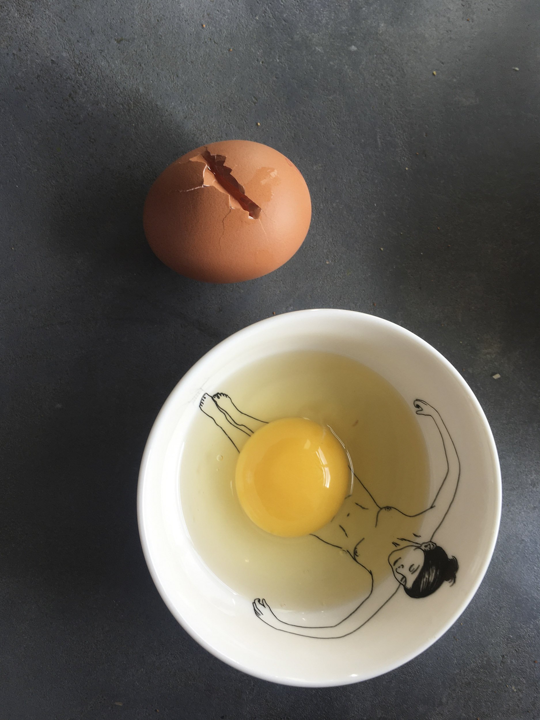 Ein rohes Ei in einer Schüssel mit einer Zeichnung eines liegenden Menschen, daneben ein weiteres Ei.