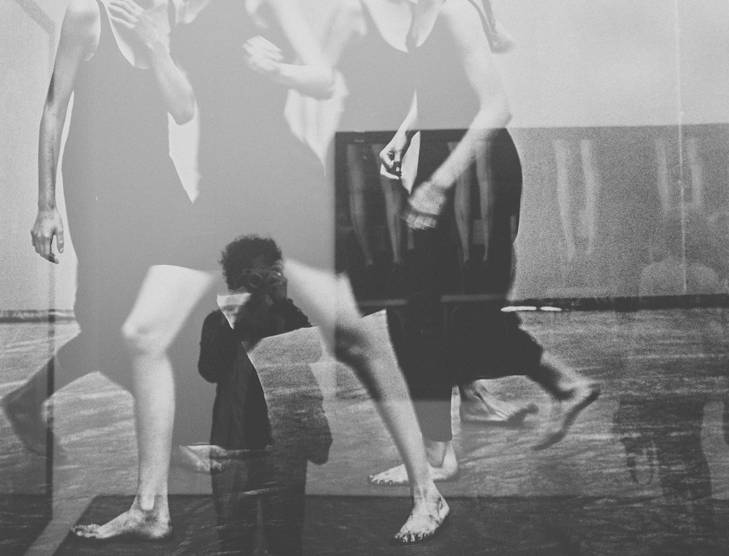 Mehrere Menschen, die barfuß Yoga oder Tanz machen, sichtbar durch Doppelbelichtung. Im Vordergrund stehen nur die Beine, im Hintergrund sind weitere Personen, inklusive einem Kind mit Brille, sichtbar, die in einem Raum üben.