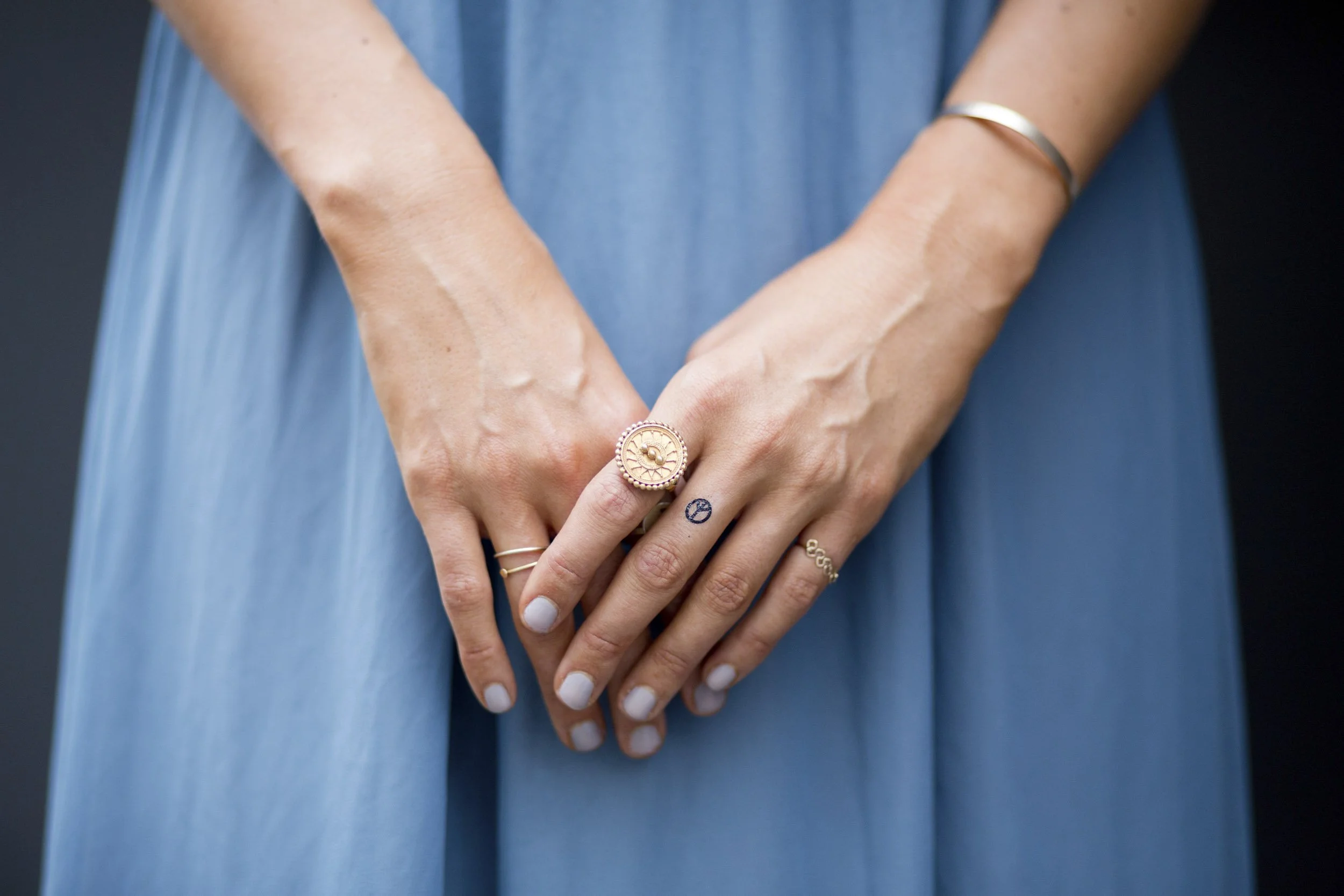 Nahaufnahme von Händen, die in einem blauen Kleid gekreuzt sind, mit mehreren Ringen und Tattoos auf den Fingern.