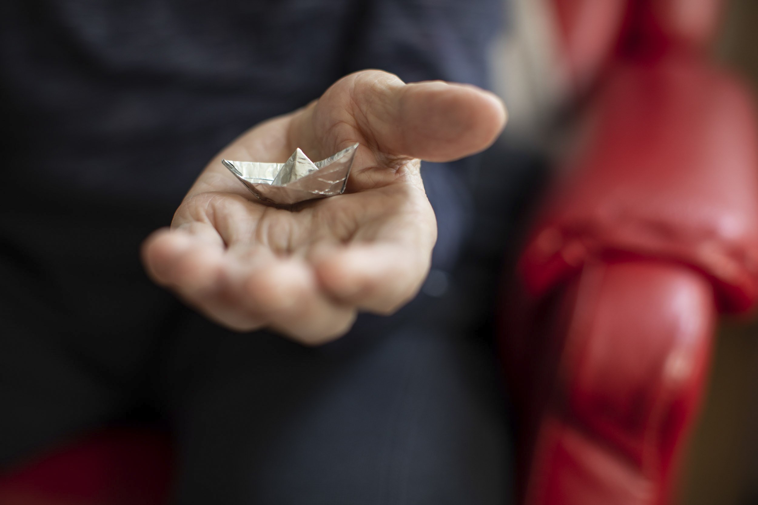 Eine Hand hält ein kleines aus Aluminiumfolie Schiff. Im Hintergrund sind rote Schuhe sichtbar.