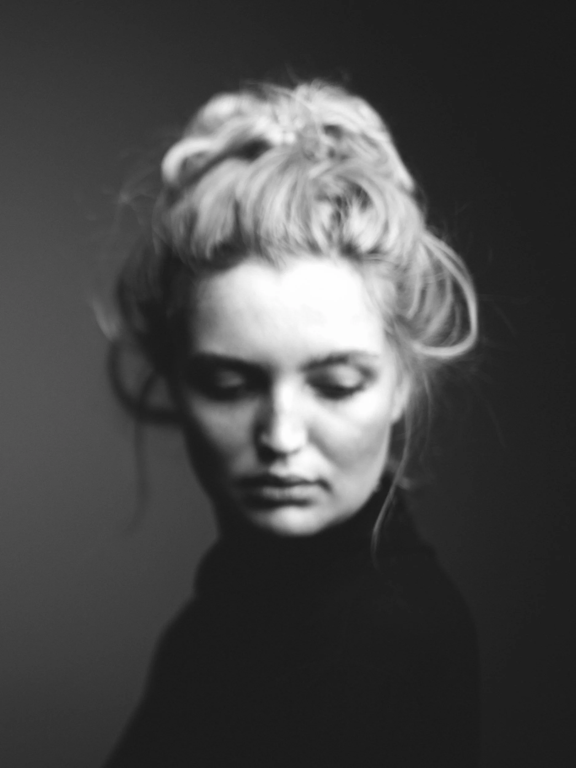 Schwarz-weiß Porträt einer Frau mit geschlossenen Augen und hochgestecktem, wolligem Haar vor dunkelgrauem Hintergrund.