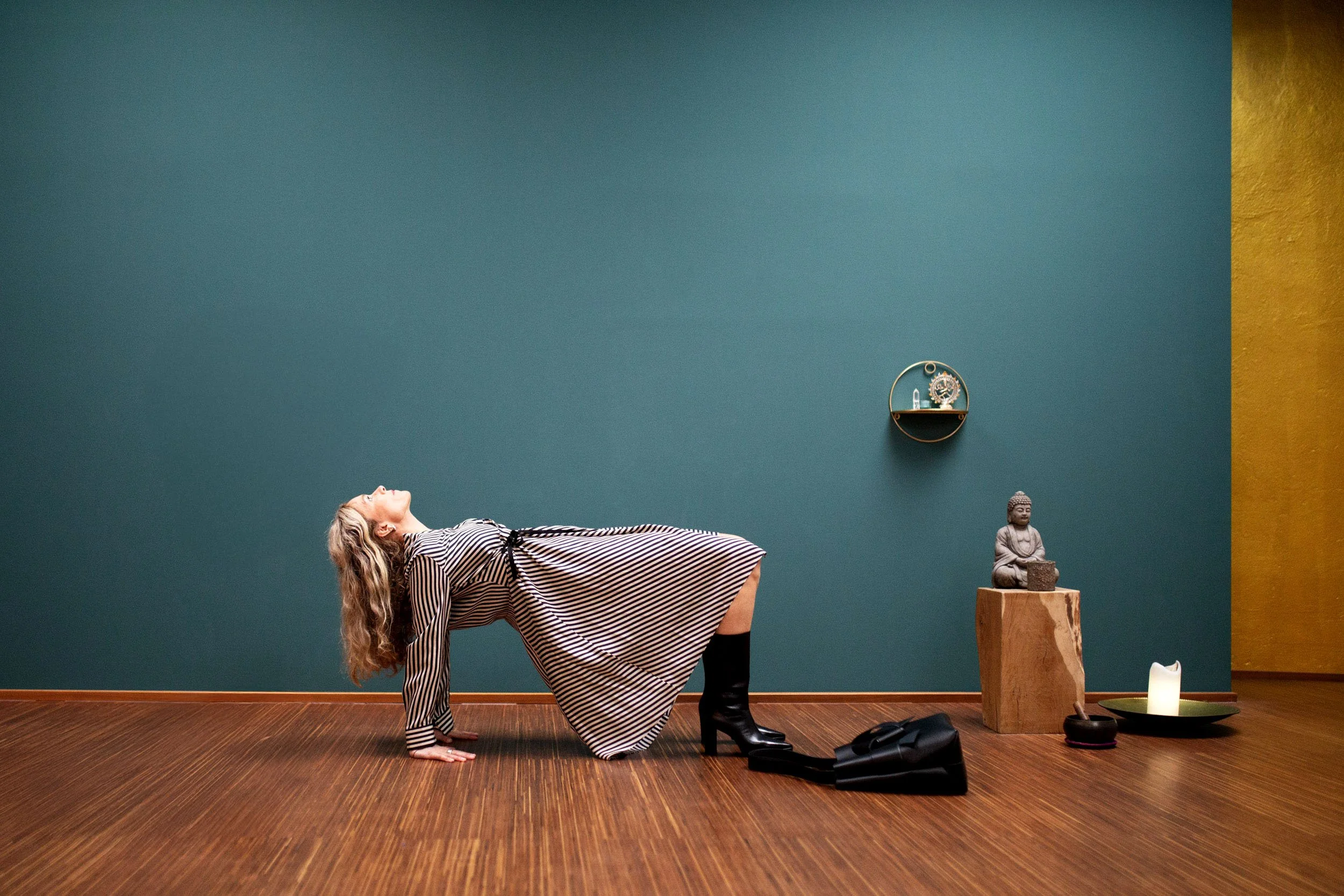 Frau in gestreiftem Kleid in einem Raum mit blauer Wand und Holzboden. Es sind Dekorationsgegenstände wie eine Buddha-Statue, Kerzen, eine kleine Uhr und weitere Gegenstände auf einem Holzstapel und kleinen Regalen sichtbar.