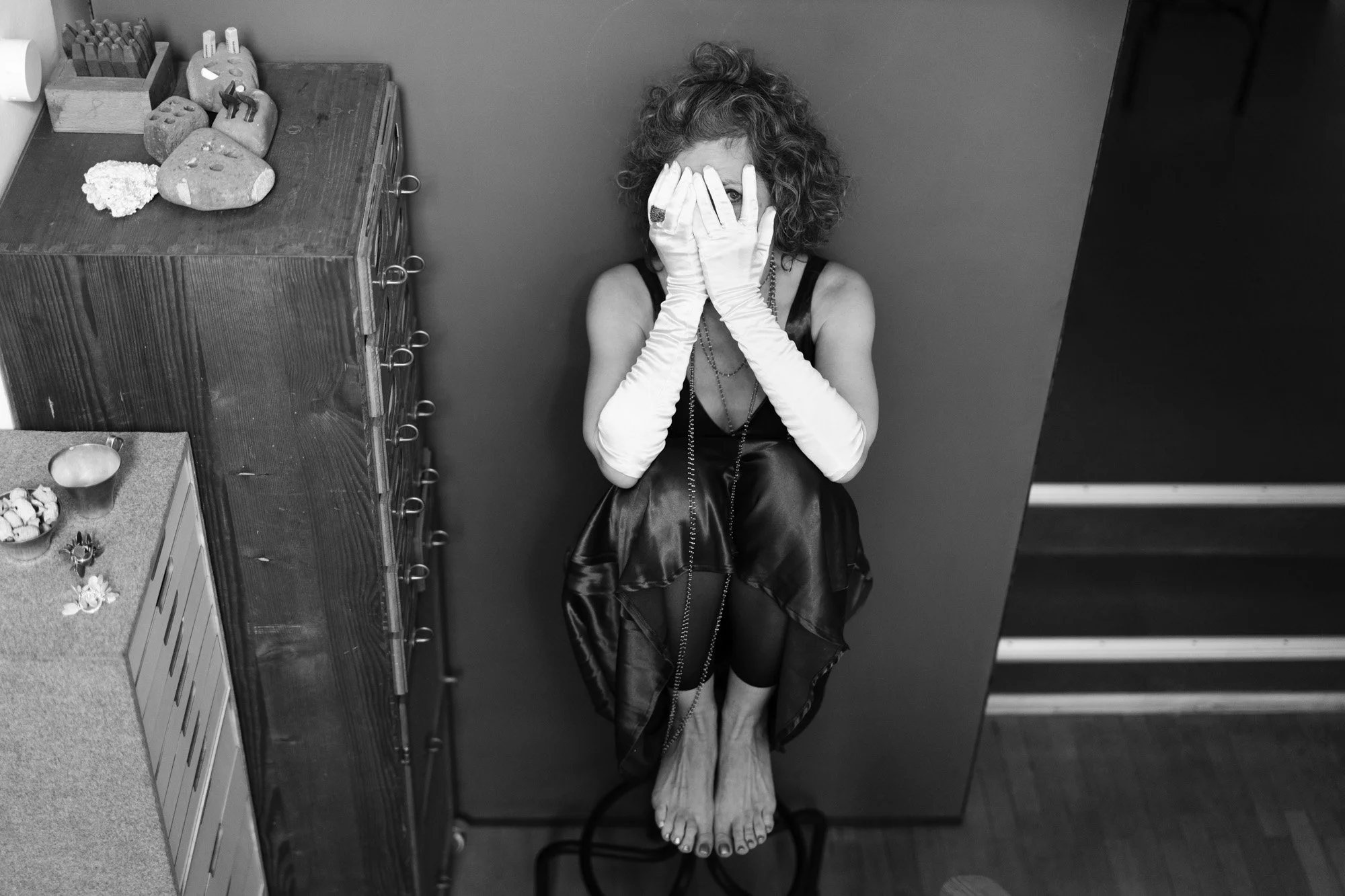Eine Frau mit zersaustem, kurzen Haar sitzt an einer Wand, die Hände vor das Gesicht haltend und nur die Augen sichtbar. Sie trägt lange weiße Handschuhe, ein schwarzes Kleid und Netzstrümpfe. Es ist ein Schwarz-Weiß-Foto.