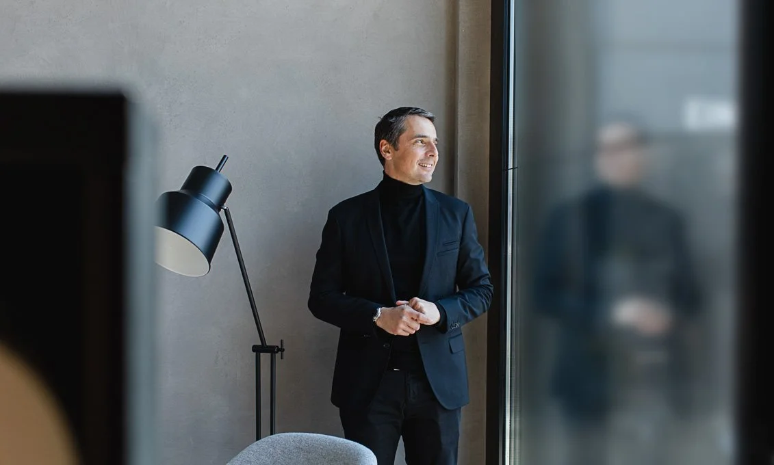 Ein Mann steht in einem modernen Büro neben einem Fenster, schaut nach draußen und lächelt.
