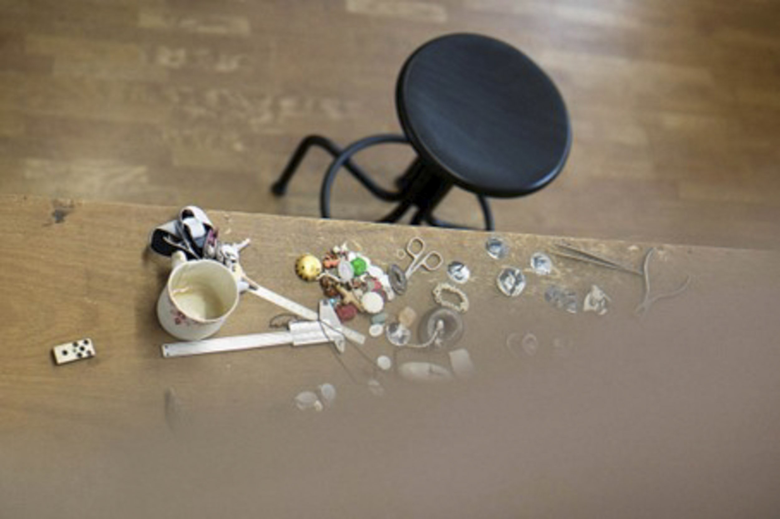 Auf einem Holztisch liegen zerstreute Gegenstände, darunter eine Tasse, kleine Spielzeugfiguren, Schmuck, eine Pinzette, Glaskristalle und ein Domino-Steinen. Im Hintergrund steht ein schwarzer Hocker.