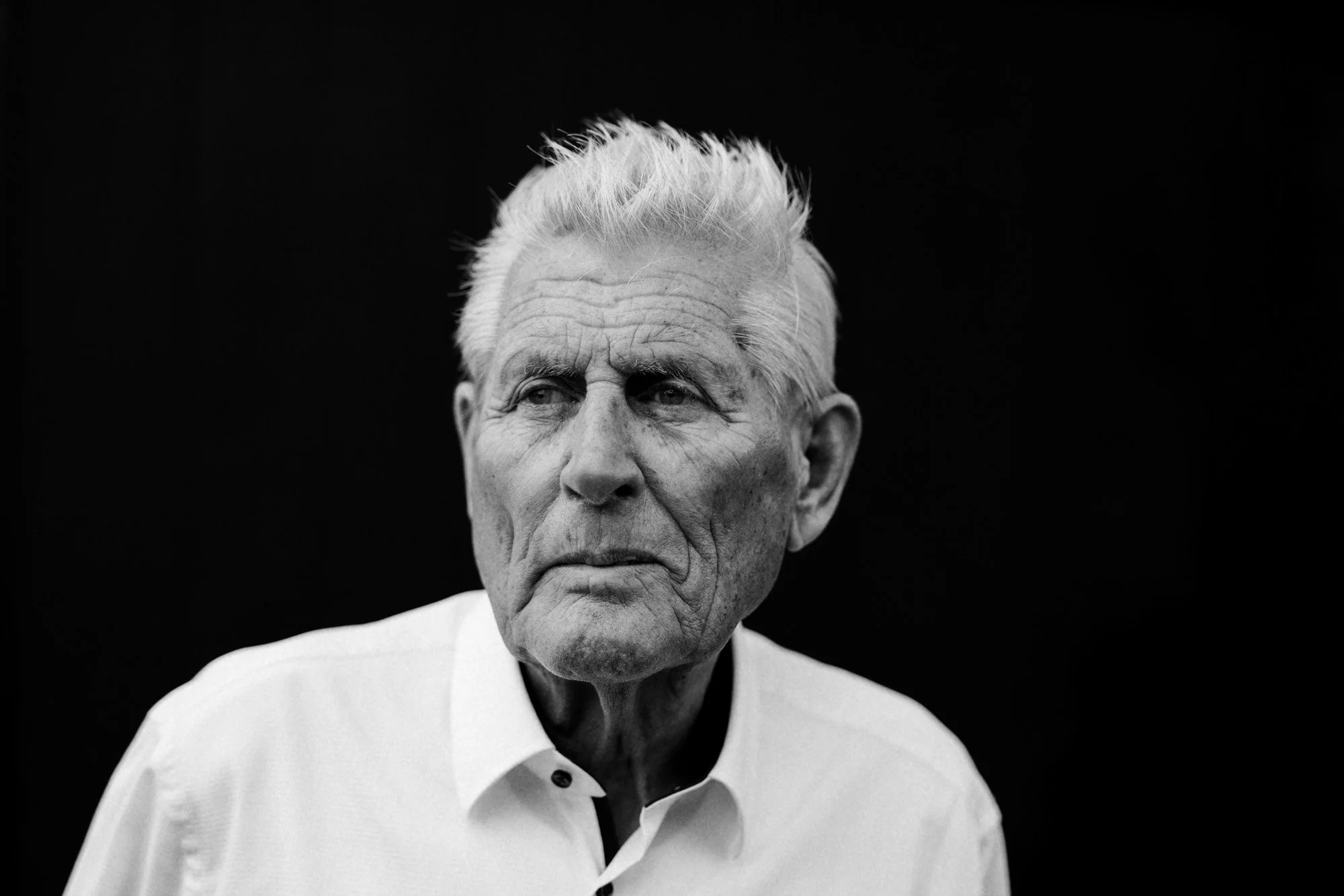 Schwarz-Weiß-Foto eines alten Mannes mit weißen Haaren, der nach links blickt, vor schwarzem Hintergrund, trägt ein Hemd.