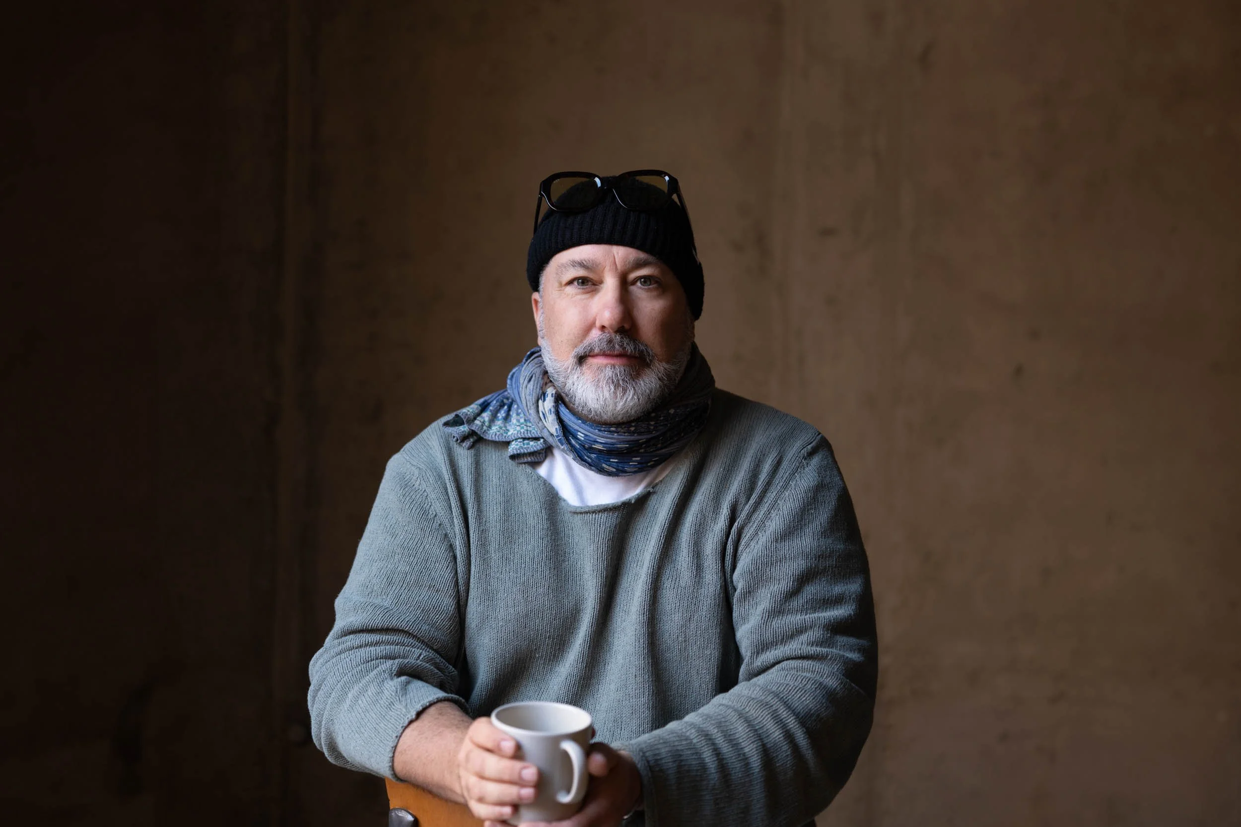 Ein Mann mit grauem Bart trägt eine schwarze Mütze, eine Brille auf dem Kopf, eine graue Jacke, einen Schal und hält eine weiße Tasse in den Händen, vor einem braunen Hintergrund.