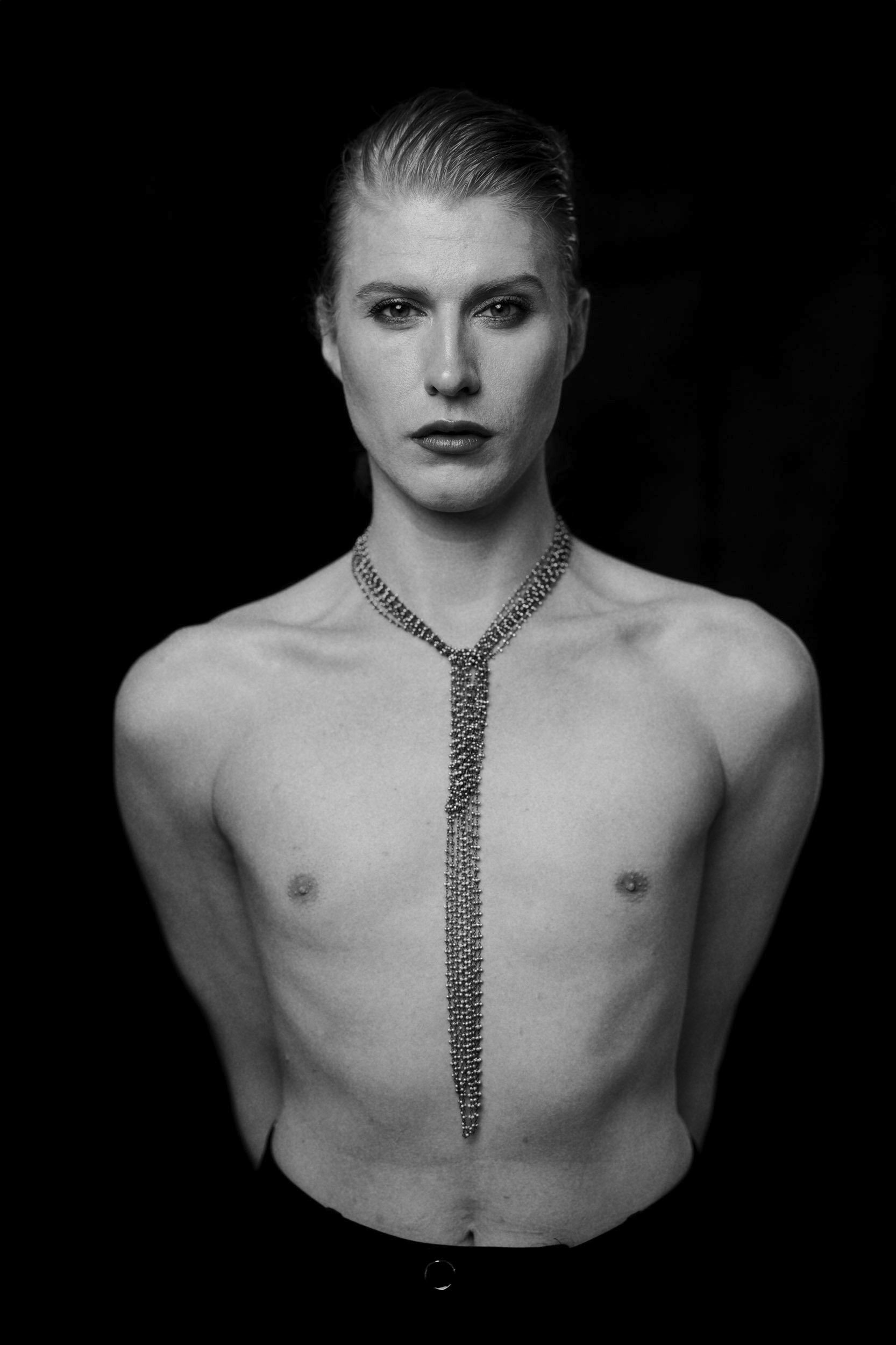 Schwarz-weiß-Fotografie eines  Mannes mit kurzen, nach hinten gekämmten Haaren, der eine lange Halskette trägt, vor schwarzem Hintergrund.