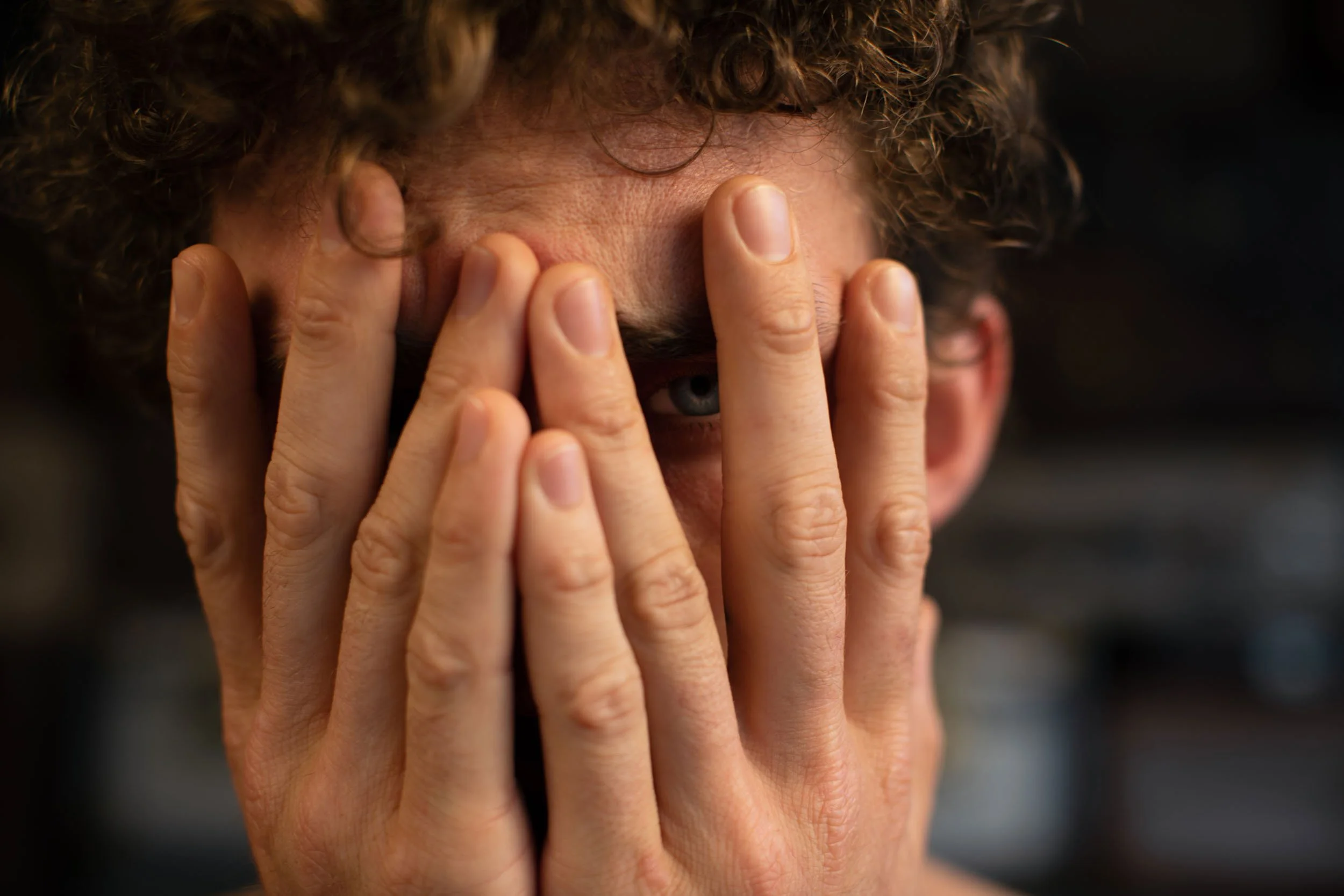 Eine Person mit lockigem Haar, die sich die Hände ins Gesicht hält, wobei nur ein Auge sichtbar ist.