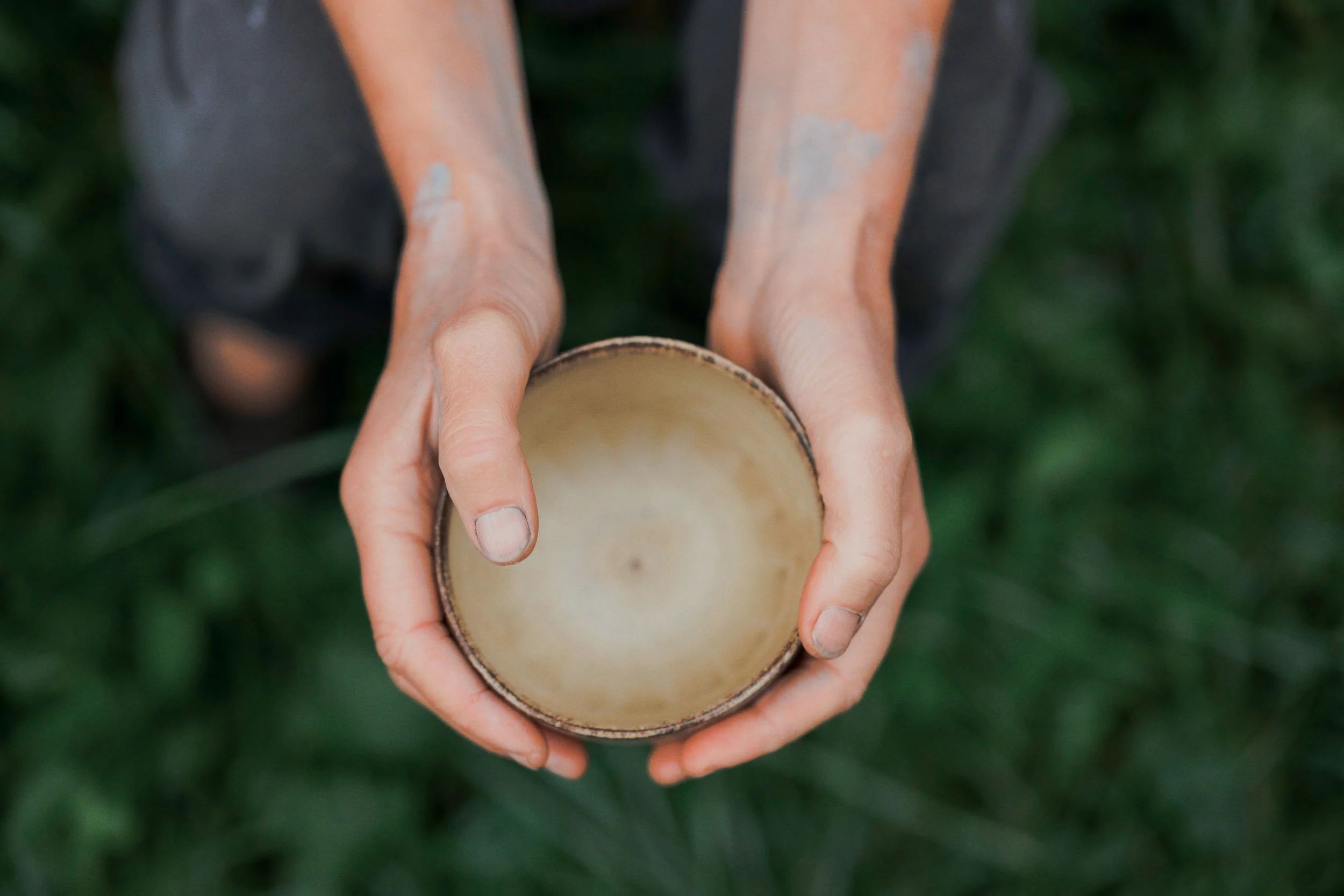 Nahaufnahme von Händen, die eine leere, runde Tasse oder Schüssel halten, vor grünem Gras, aus der Vogelperspektive.