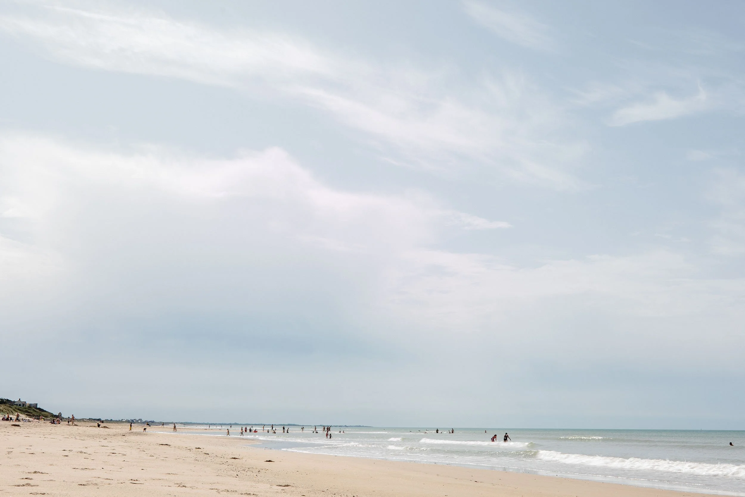 Strand mit wenigen Menschen im Meer und am Strand, Wolkenhimmel, Küstenlinie im Hintergrund.