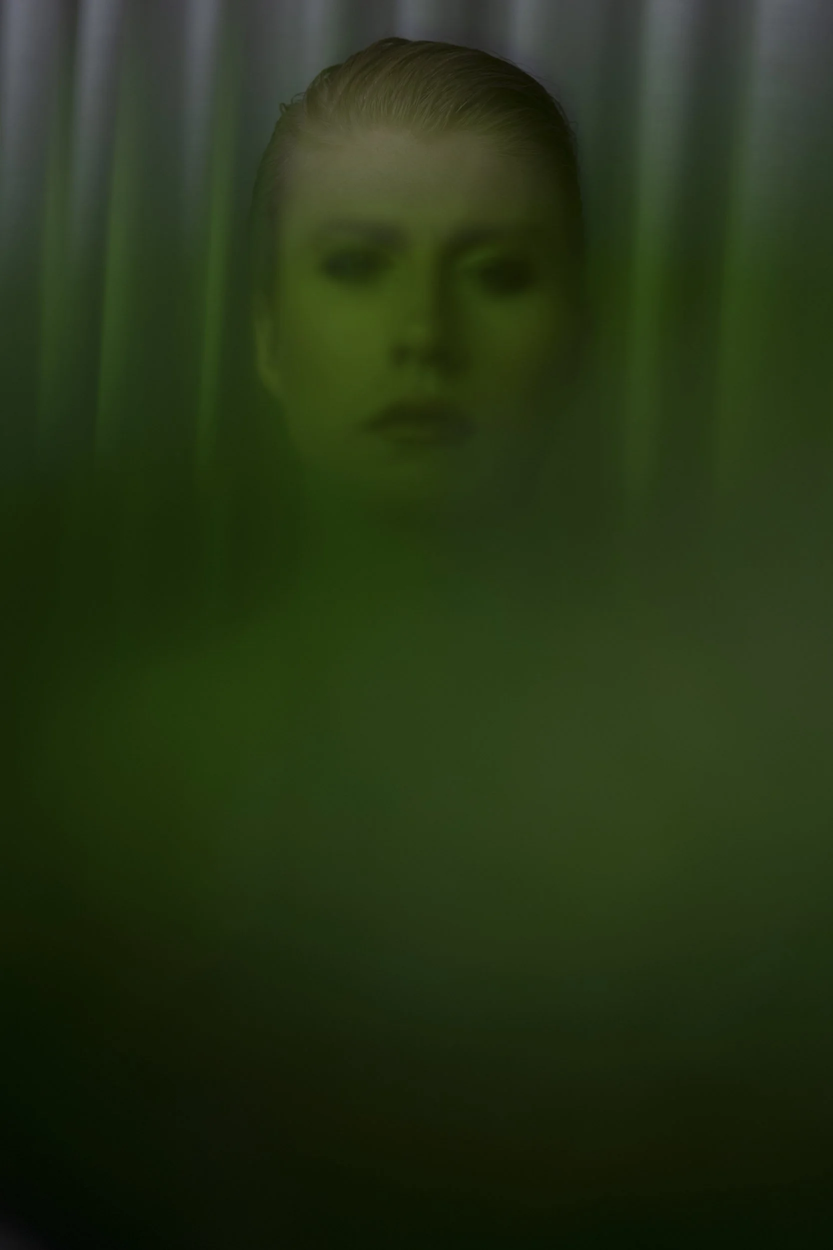 Porträt einer jungen Frau, teilweise verdeckt durch grüne Vorhänge, mit glatter, nasser Frisur und ernstem Gesichtsausdruck.