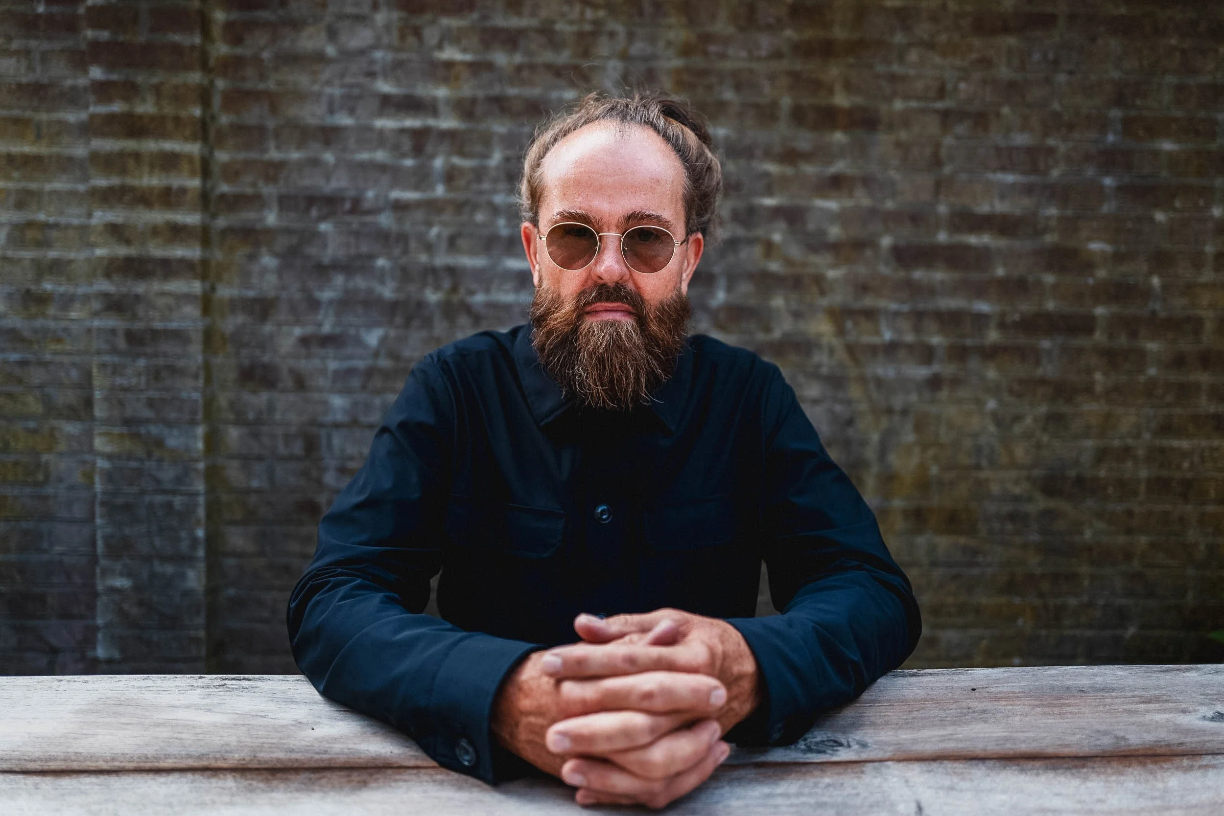 Ein Mann mit Bart, Sonnenbrille und dunklem Hemd sitzt an einem Holztisch vor einer Backsteinmauer.