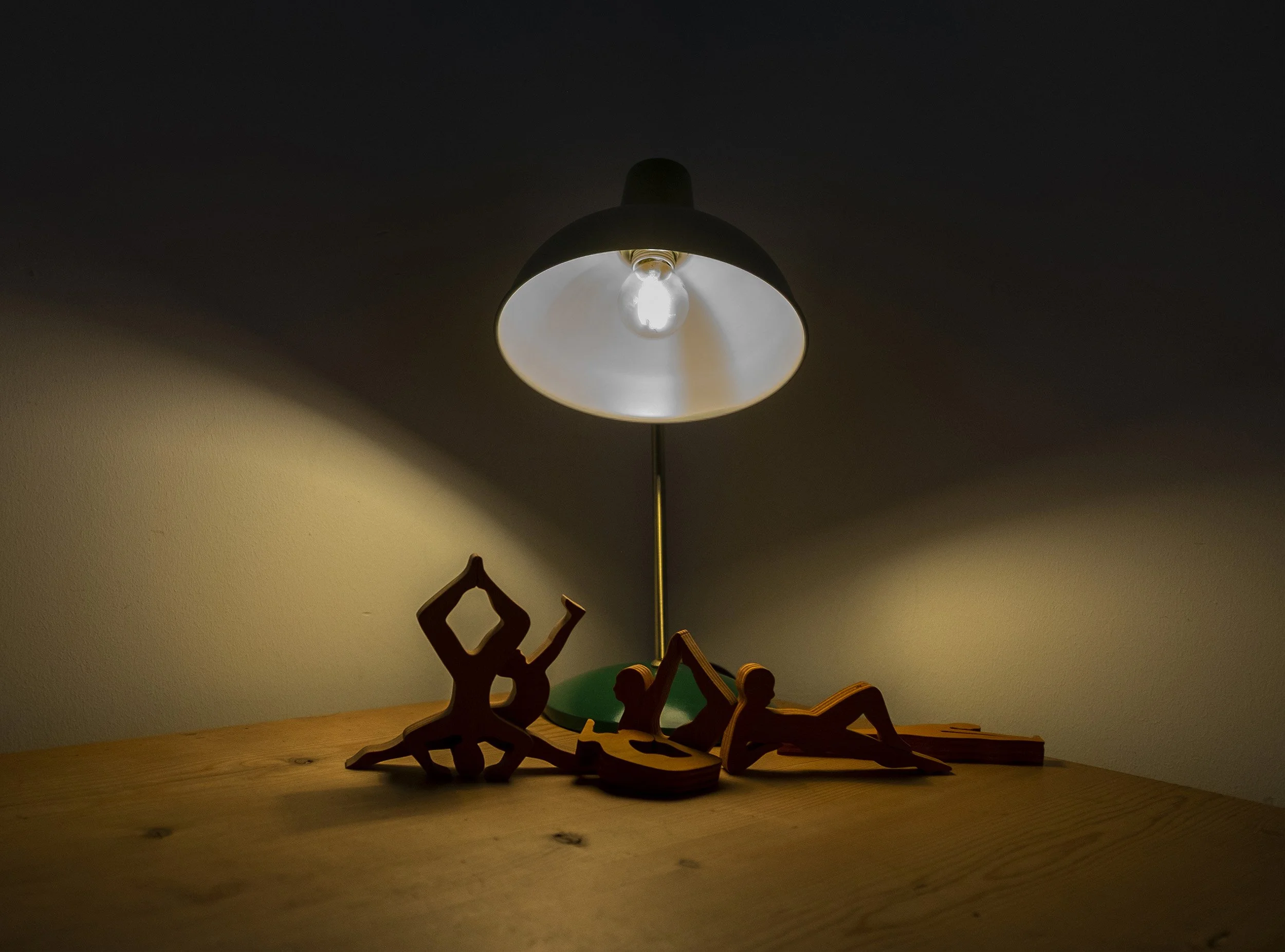 Holzskulpturen von Yogafiguren vor einer Schreibtischlampe auf einem Holztisch, mit Licht und Schatten im Raum.
