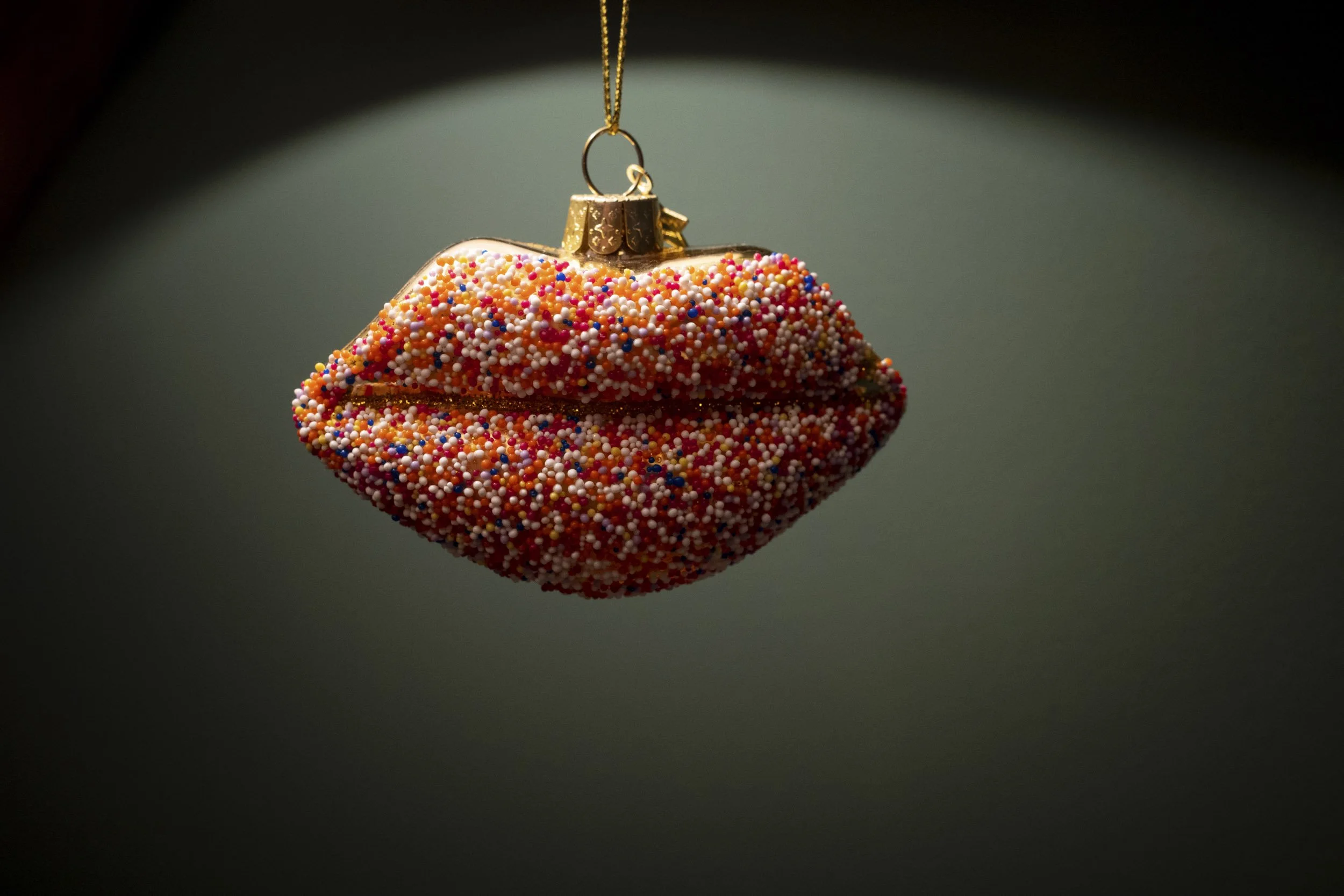 Eine Glasornament in Form von Lippen, bedeckt mit bunten Streuseln, hängt an einer goldenen Kette.