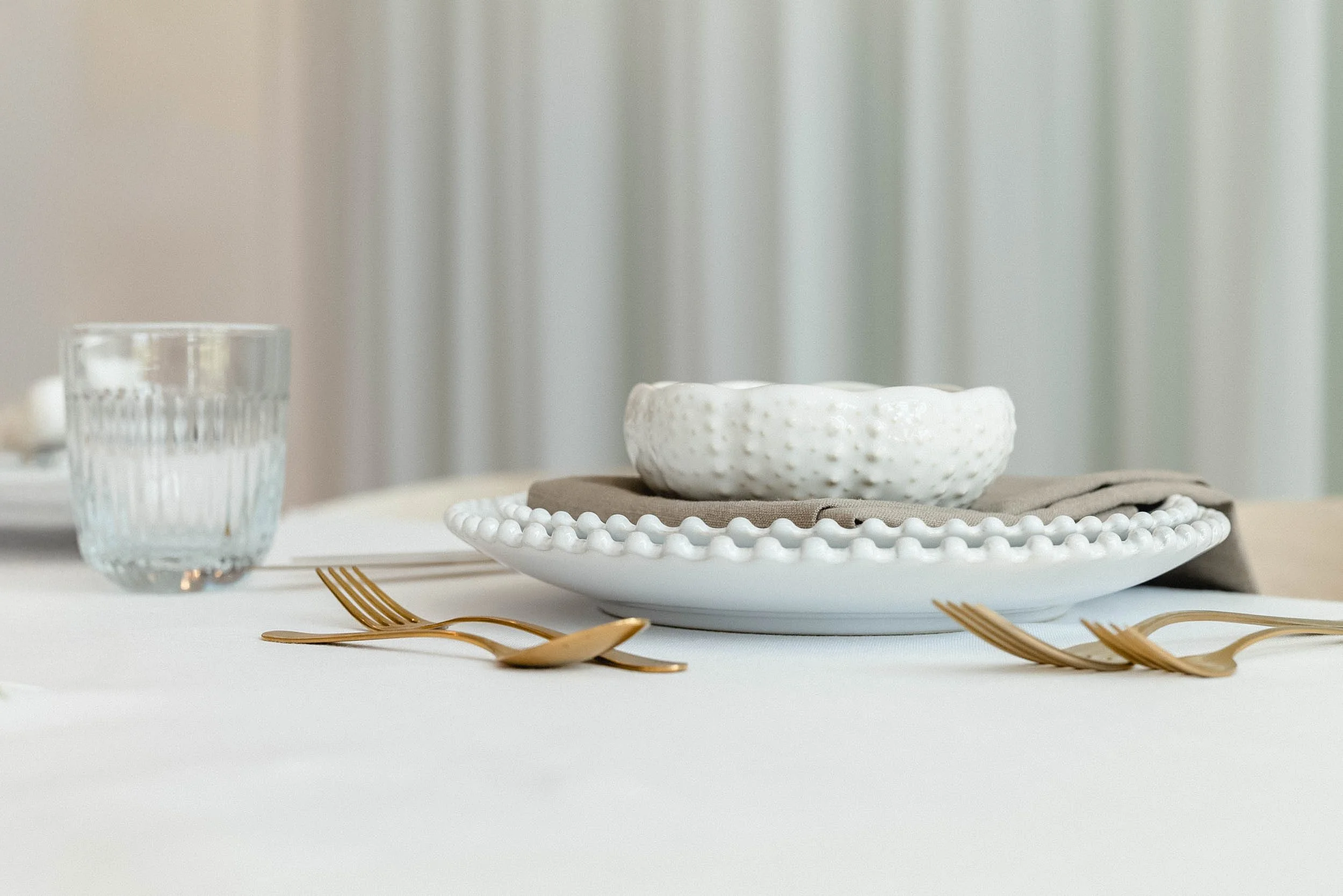 Ein gedeckter Esstisch mit weißem Teller, einer weißen Schüssel mit dicker Struktur, einer beige Serviette, einer Wasser glass und goldfarbenen Besteck. Im Hintergrund sind verwaschene Vorhänge.