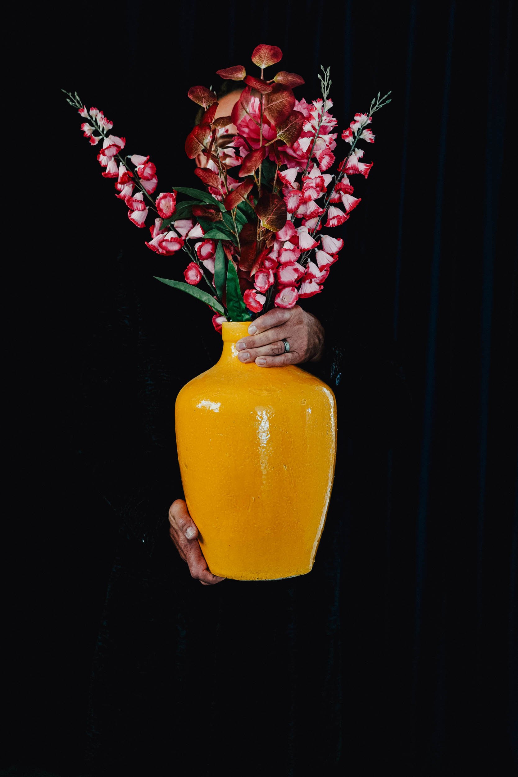Eine Person hält eine große, gelbe Vase mit roten und weißen Blumen gegen einen dunklen Hintergrund.