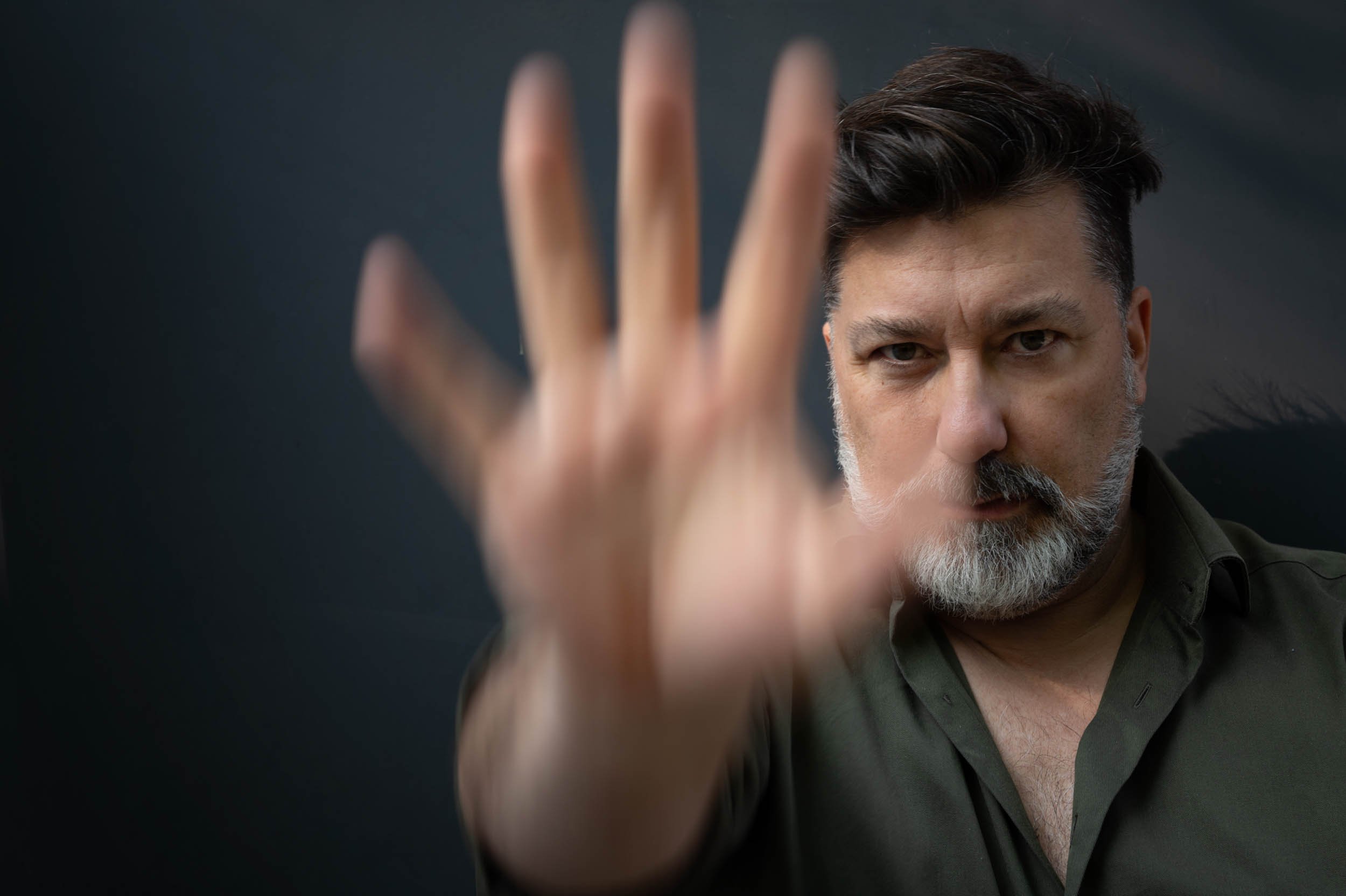 Ein Mann mit dunklem Haar und grau-weißem Bart zeigt mit seiner Hand nach vorne, während er ernst schaut, vor einem dunklen Hintergrund.