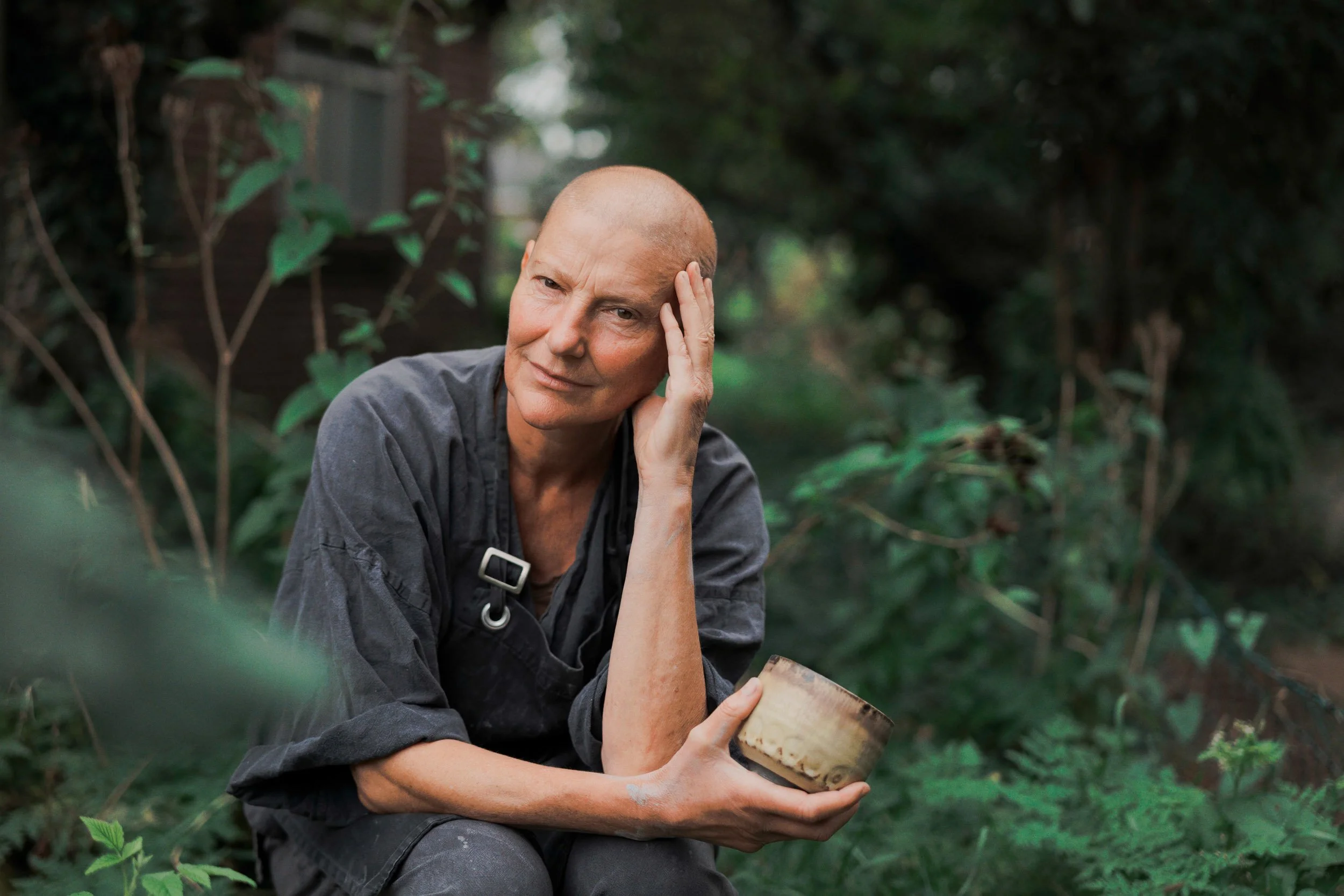 Künstlerin im Garten, die eine Keramik hält, während sie nachdenkt, mit einem neutralen Ausdruck auf dem Gesicht.