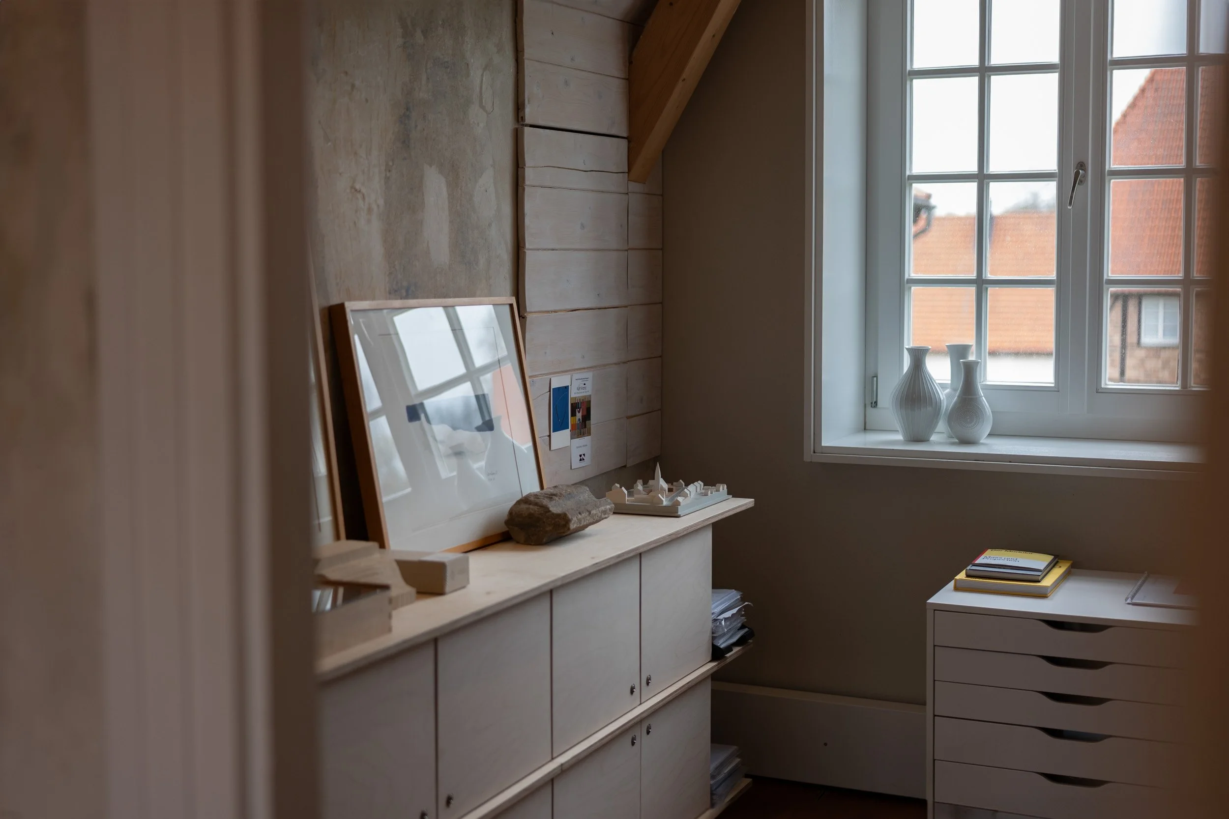 Ein heller Raum mit einem Fenster, auf der Fensterbank stehen zwei weiße Vasen, und auf einem weißen Schrank liegen Bücher und Papiere, im Hintergrund eine ballaststoffreiche Wand und ein Bilderrahmen.