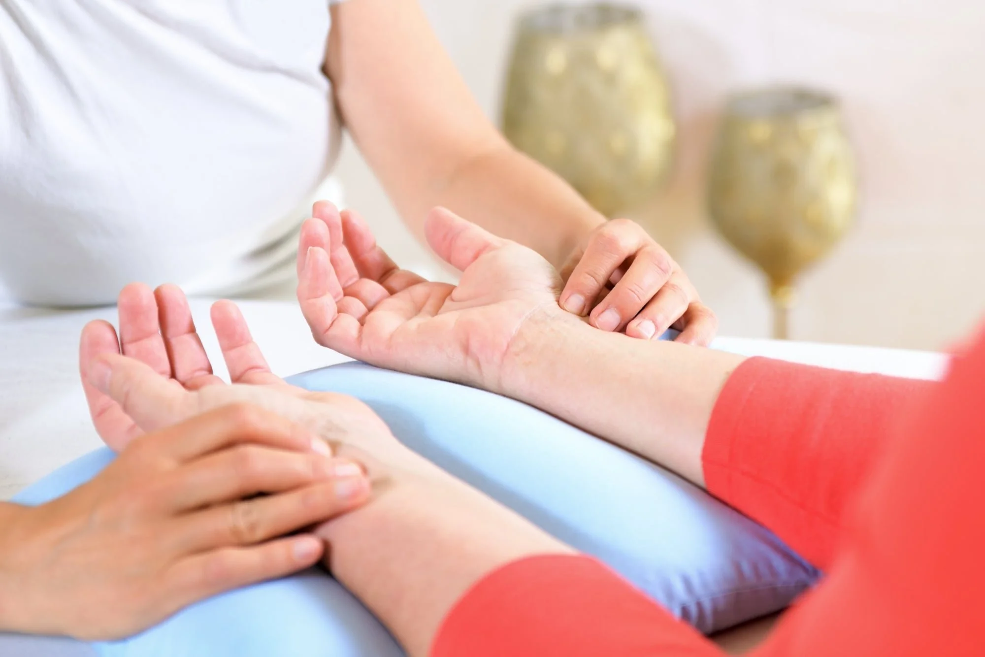 Eine Person erhält eine Handmassage bei einer Therapie, während eine andere Person ihren Arm massiert. Der Hintergrund ist unscharf mit goldfarbenen Dekorationen.