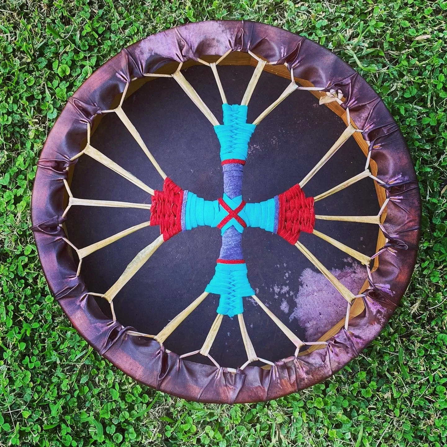 A decorative circular object resembling a dreamcatcher, made with sticks wrapped in blue and red string, stitched together with crisscrossing thread, and surrounded by a purple cloth border, placed on a green grassy ground.