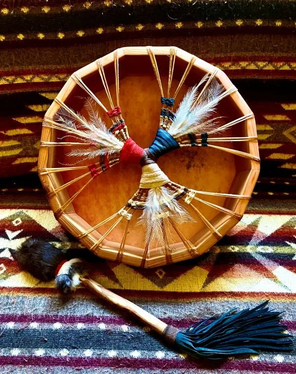A traditional Native American drum with feather decorations and a beaded stick, placed on a colorful woven rug.