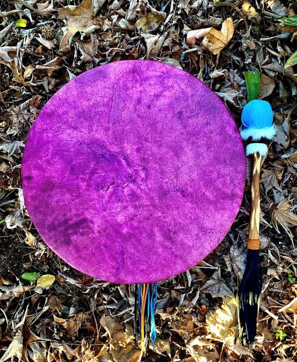 A large round purple drum with a textured surface, lying on a bed of brown leaves and twigs. There is a stick with a decorative top, featuring blue, black, and white feathers, to the right of the drum.