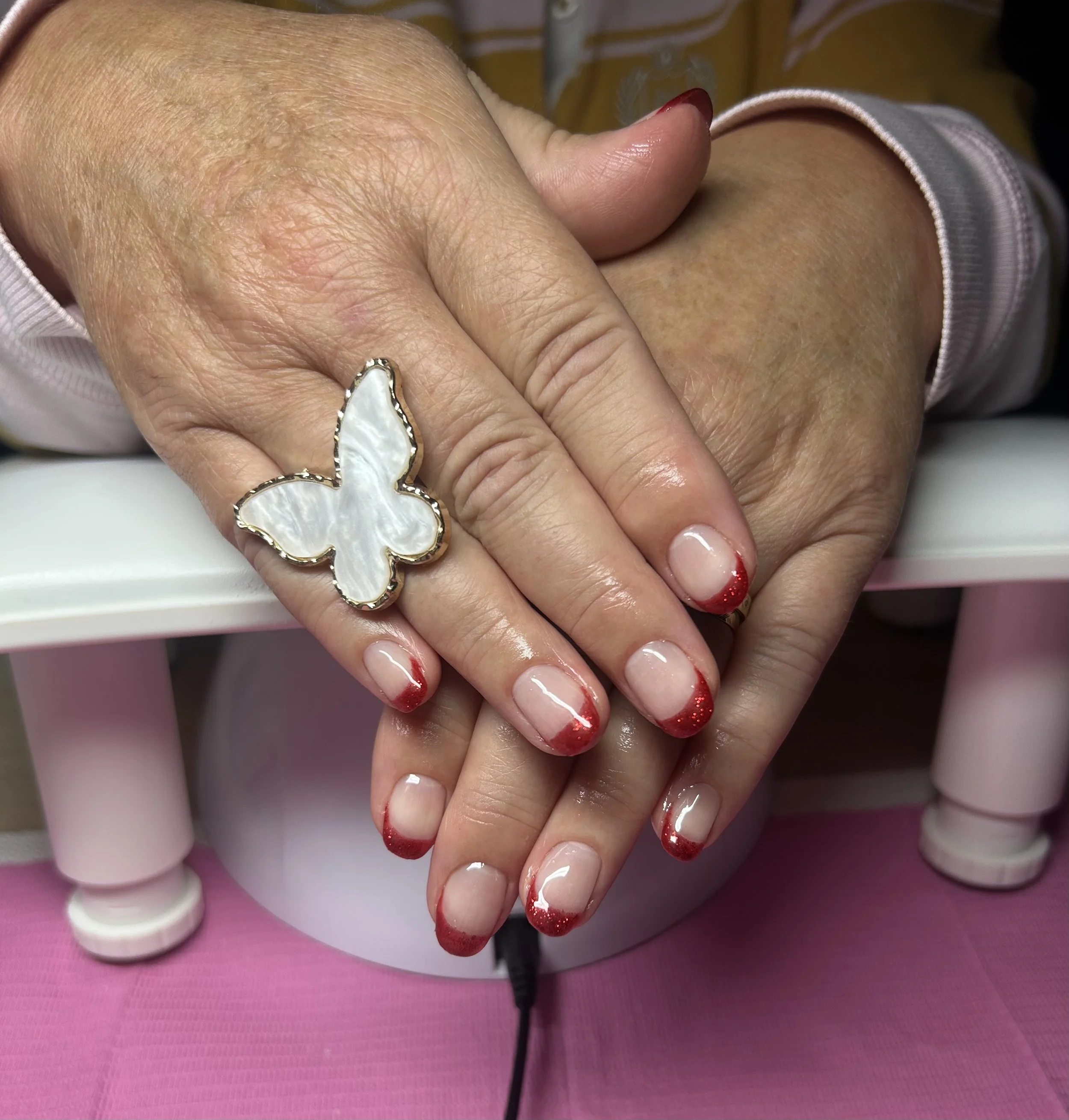 Close-up of hands with manicured nails featuring red tips, resting on a pink surface near a nail dryer. One hand has a large butterfly-shaped ring with a white, iridescent feather design.