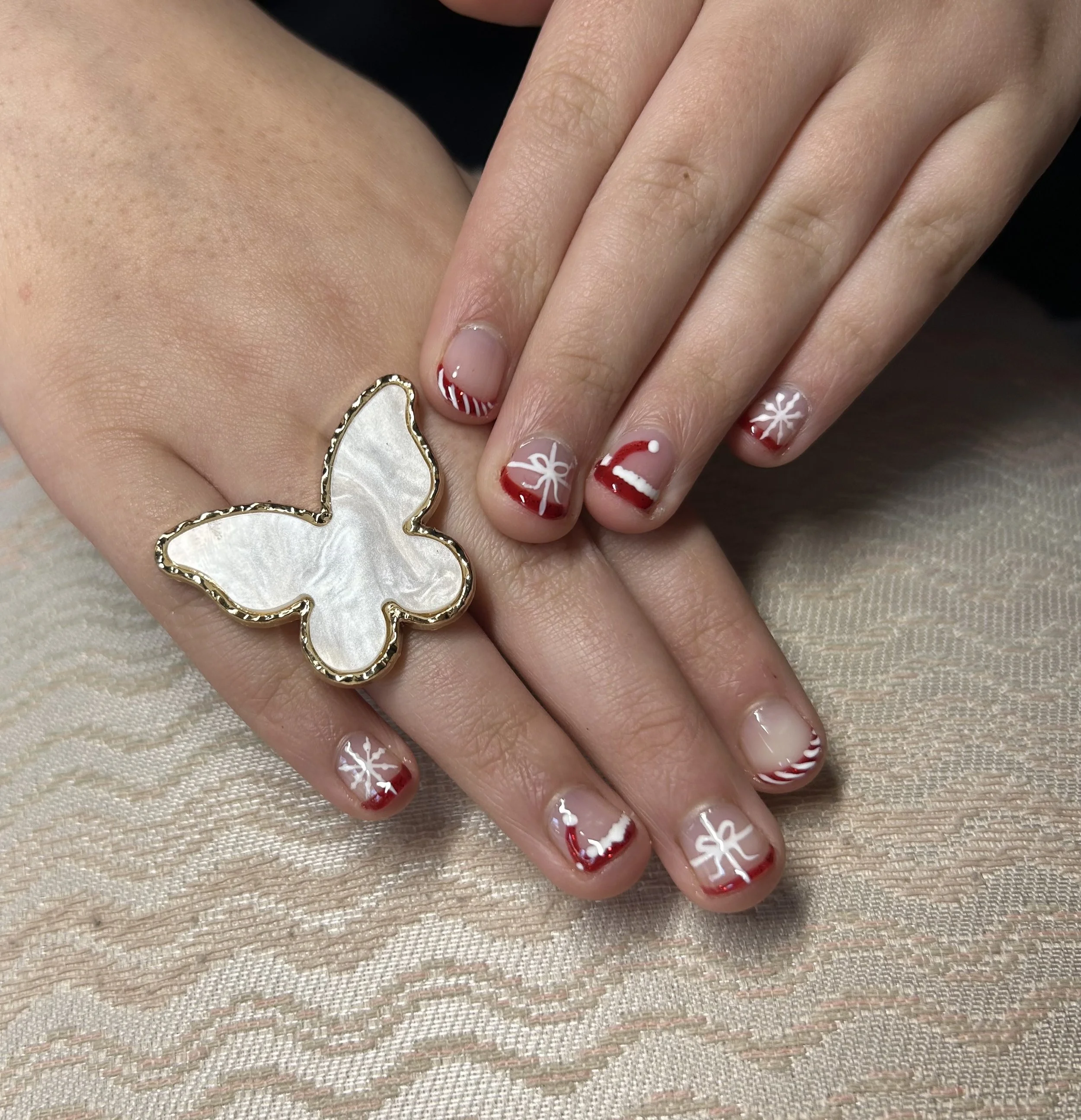 Person's hands displaying nails with red and white Christmas-themed nail art, and wearing a butterfly-shaped ring with a mother-of-pearl inlay.