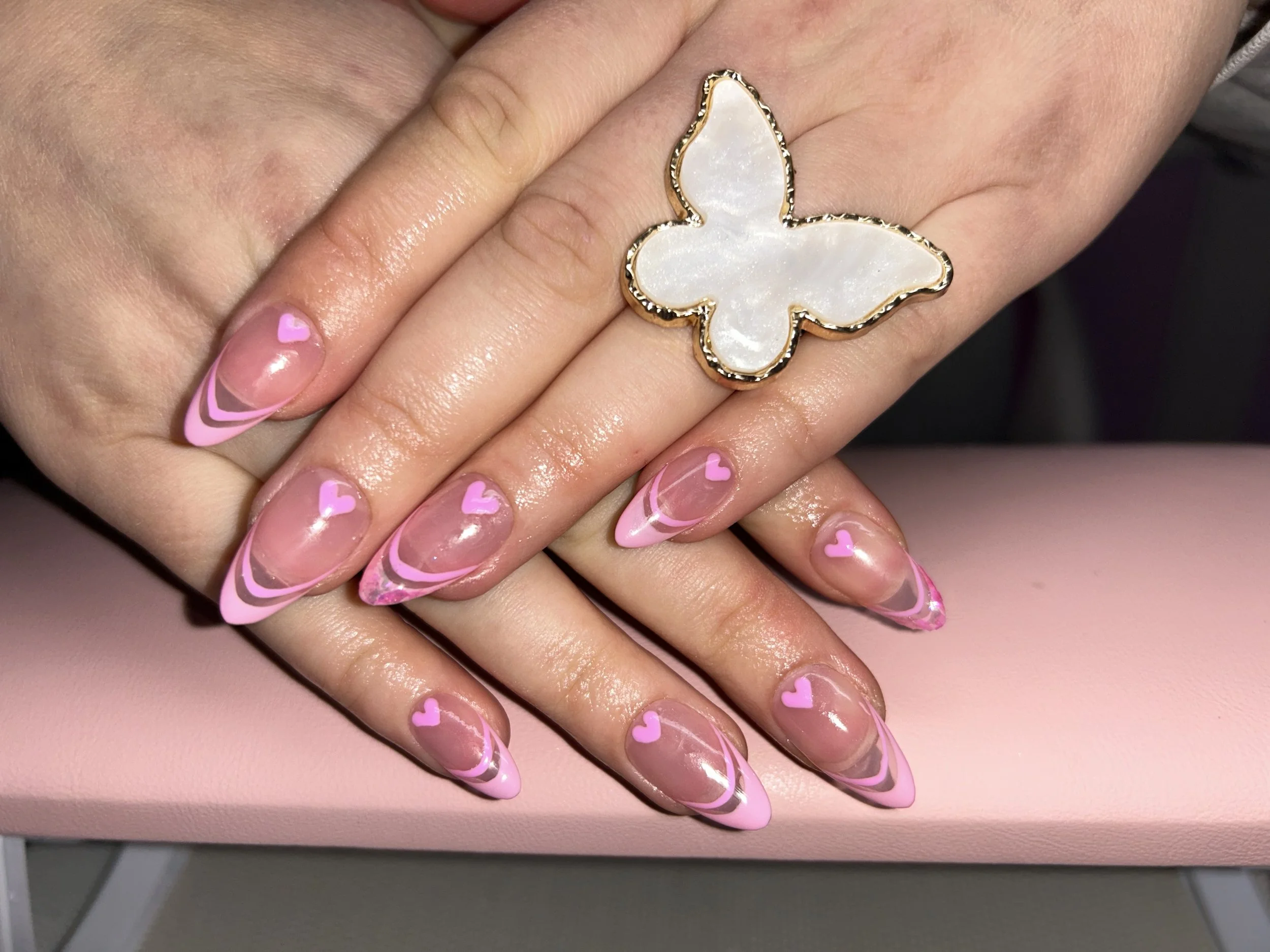 Close-up of hands showing manicured nails with pink and clear swirls and small pink hearts, and a butterfly-shaped ring with a pearl inlay on the finger.