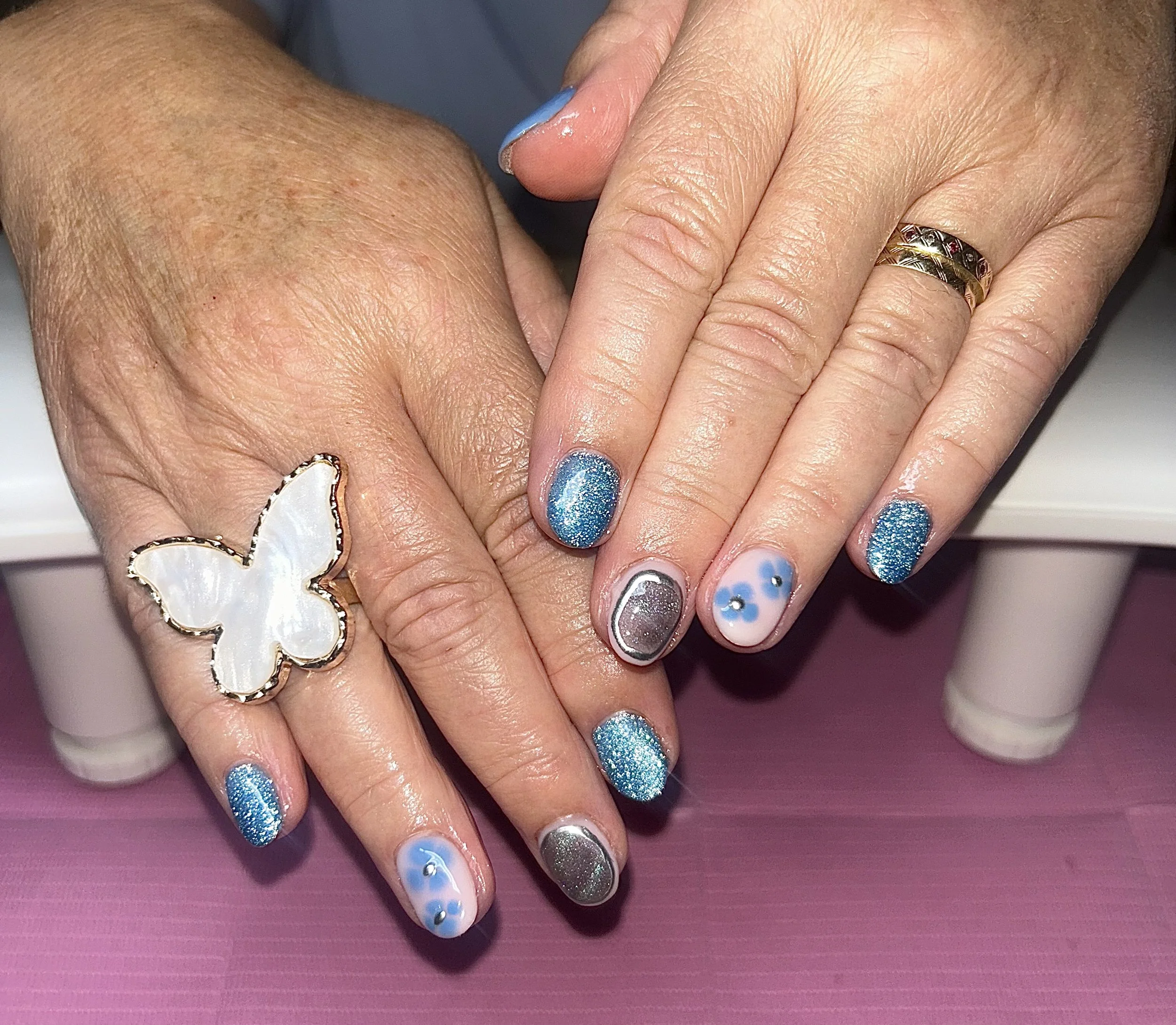 Close-up of hands with manicured nails, some painted blue with glitter, one with silver glitter, and another with a blue and white design with small rhinestones. One hand has a large butterfly ring, and the other has a gold ring with gemstones.