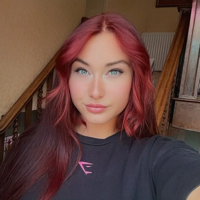 A young woman with bright red hair and blue eyes taking a selfie inside a home, with wooden stair railings and a beige wall in the background.