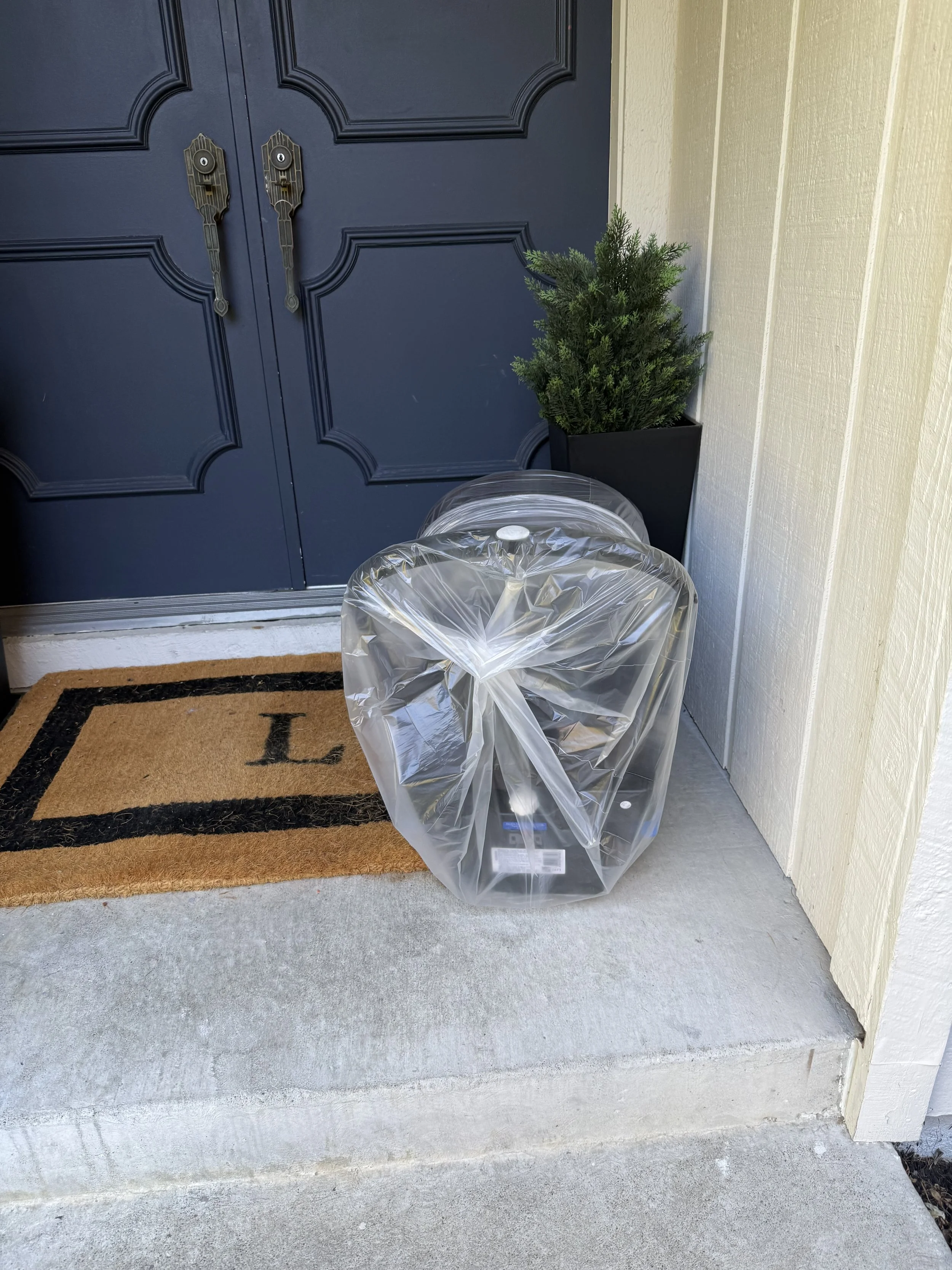 An outdoor porch area with a blue door, a small potted evergreen shrub on the right, a doormat with the letter 'L', and a plastic-wrapped car seat in front of the door.