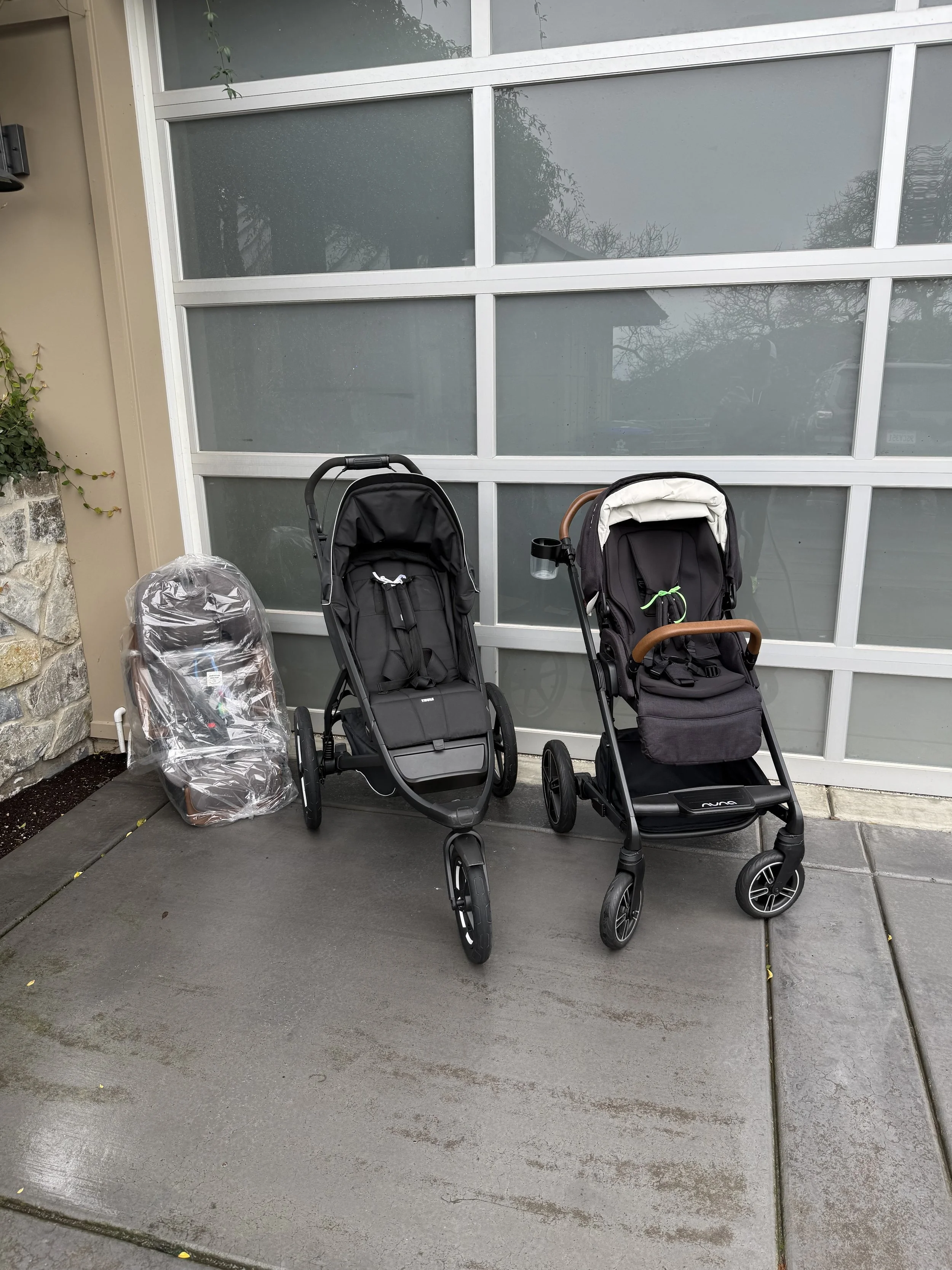 Two black strollers are parked outside in front of a glass garage door, with a plastic-covered car seat to the left and some greenery on the left side of the image.