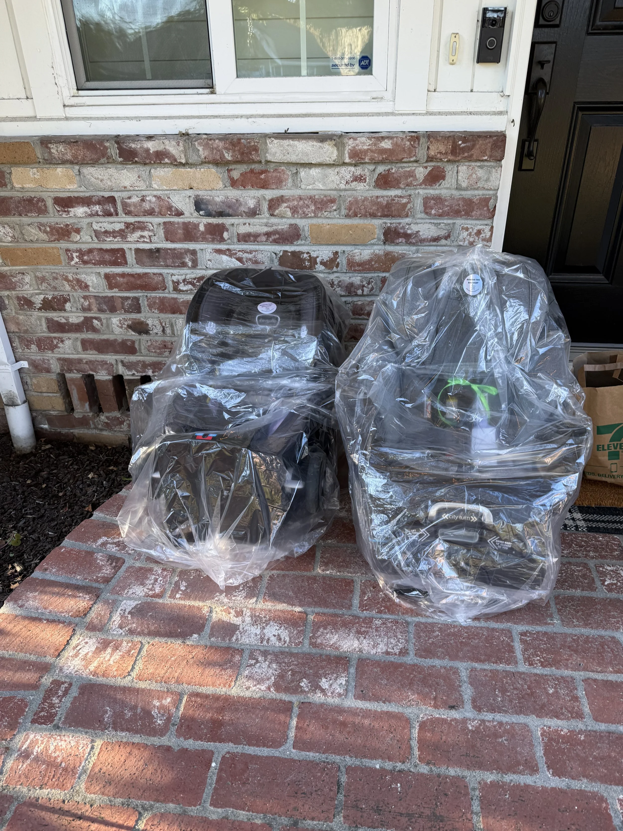 Three car seats covered with plastic bags are placed on a brick porch outside a house, near a brick wall and a black door.