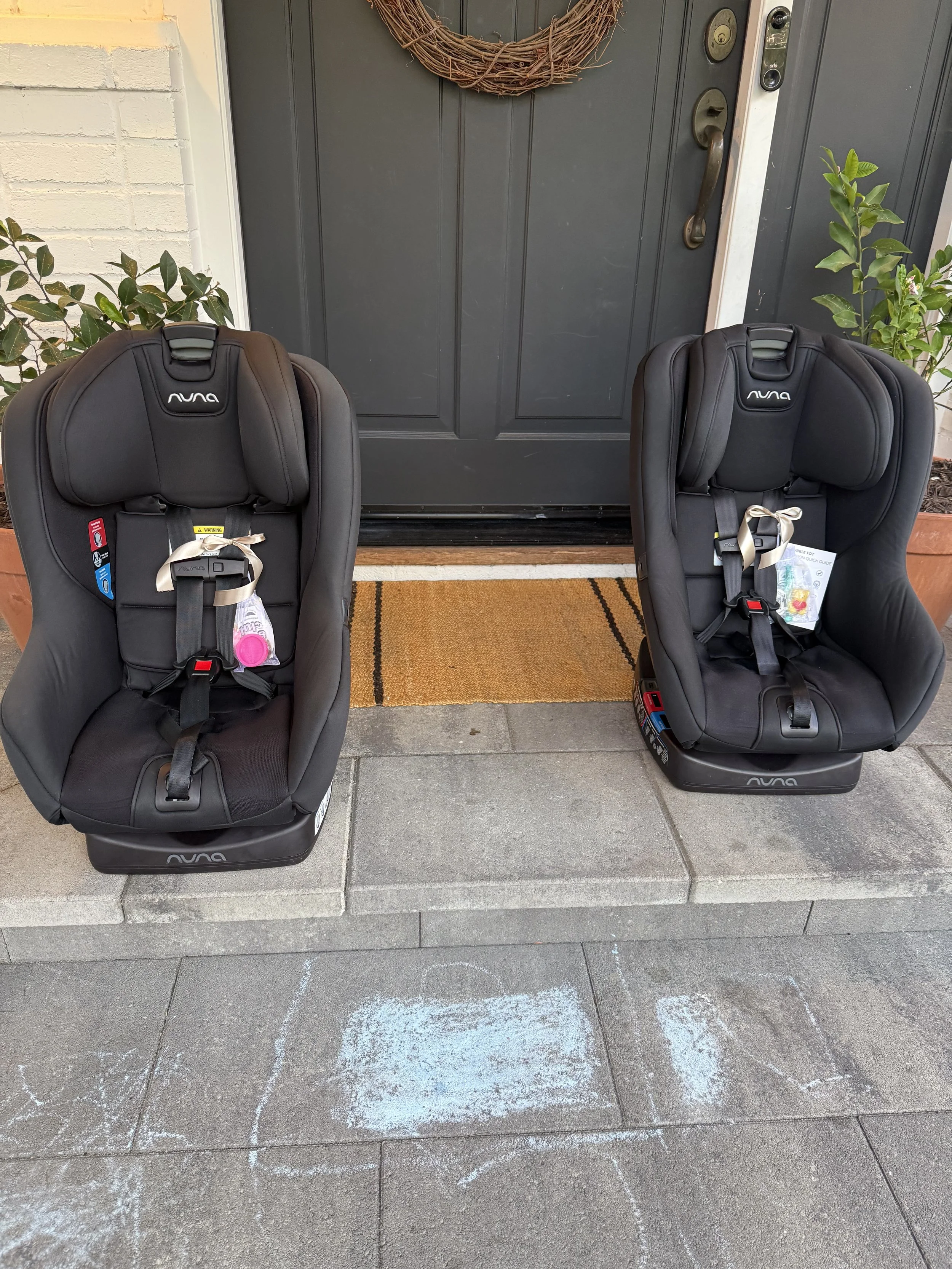 Two black Nuna car seats placed outside in front of a dark-colored door, with a doormat and sidewalk chalk drawings on the concrete underneath.