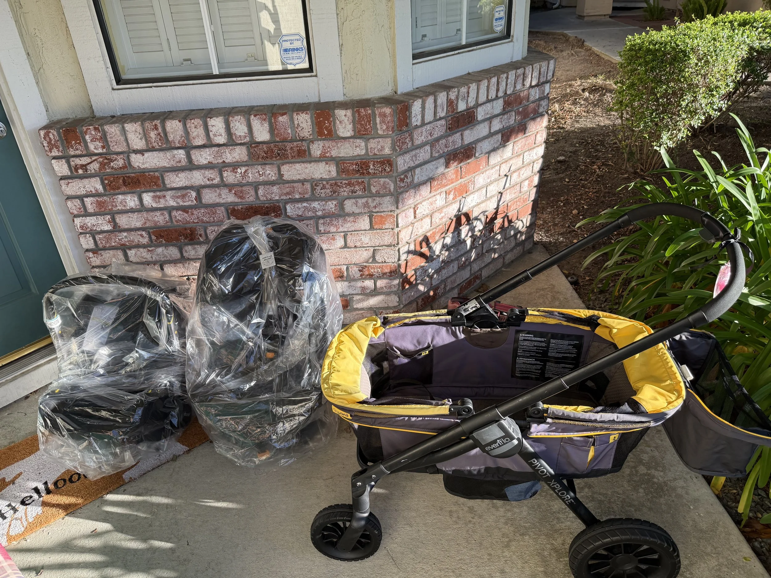 A front porch with a brick planter box, two car seats, and a yellow and gray baby wagon. There is a house door on the left and a window above the brick planter. To the right, there is a shrub and some green plants.